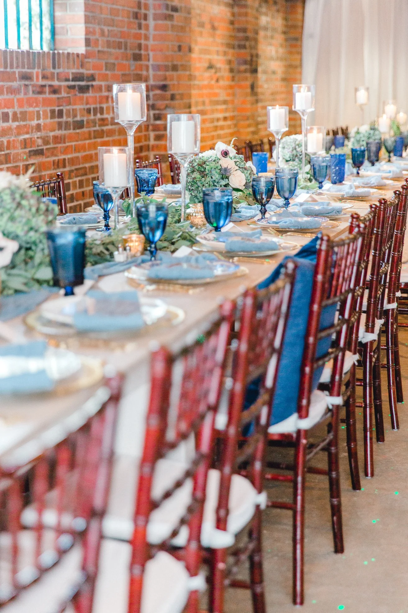 Elegant banquet table setup with blue and white dishes, blue glasses, white candles in tall holders, floral centerpieces with white roses and greenery, set against a brick wall.