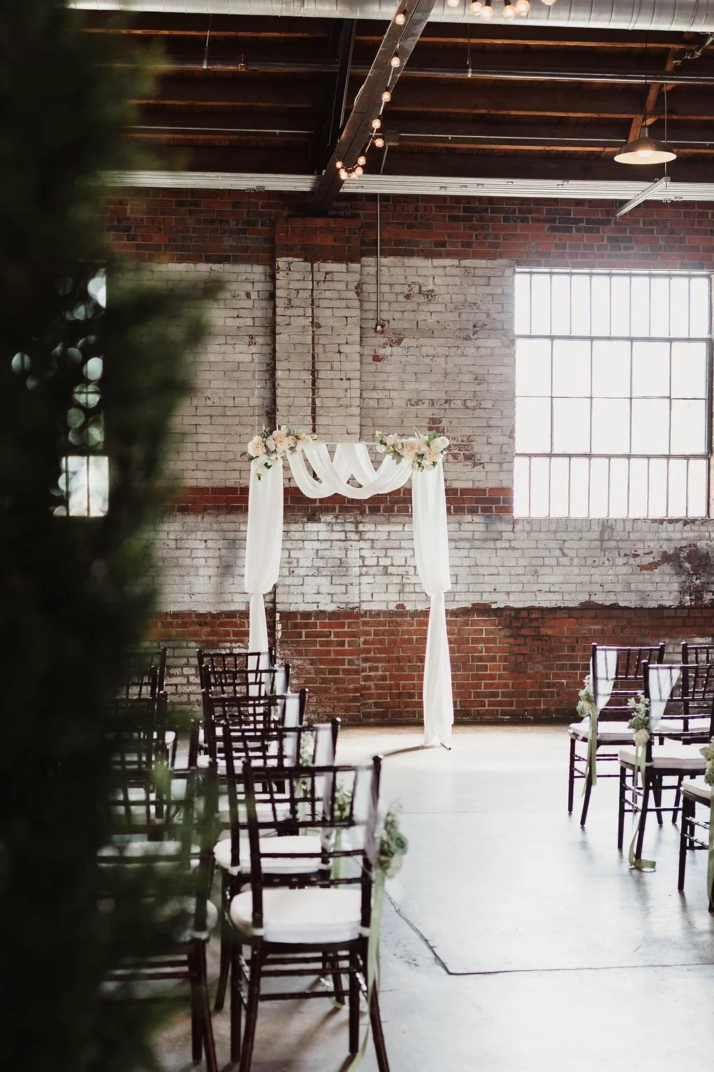Wedding aisle with chairs on both sides, decorated with flowers, leading to a white draped arch with flowers at the top, inside an industrial-style venue with exposed brick walls and large windows.