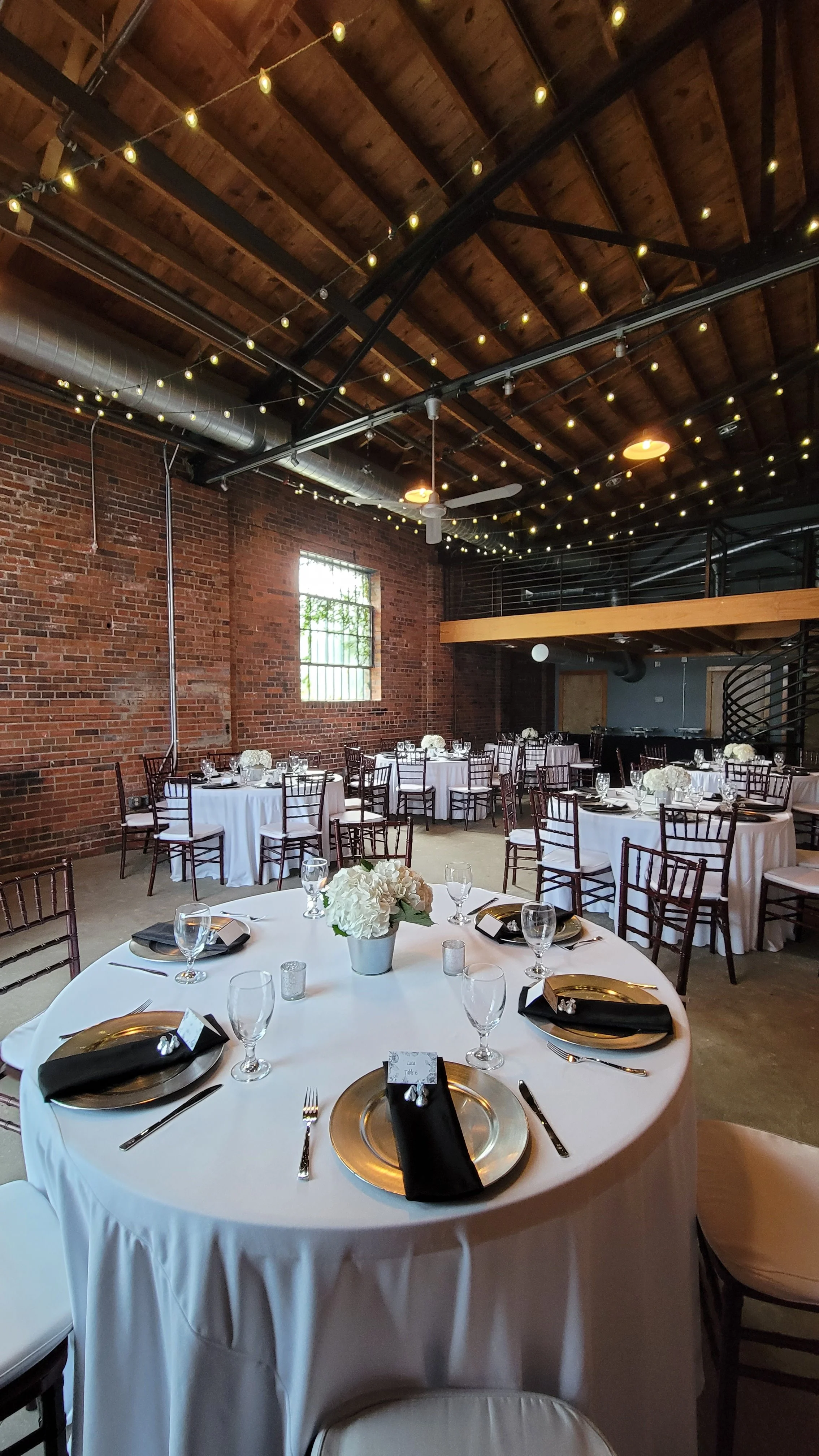 Indoor event space with round tables covered in white tablecloths, decorated with white floral centerpieces. The room features exposed brick walls, string lights hanging from the wooden ceiling, and wooden chairs around the tables. There is a second level with a railing and spiral staircase.