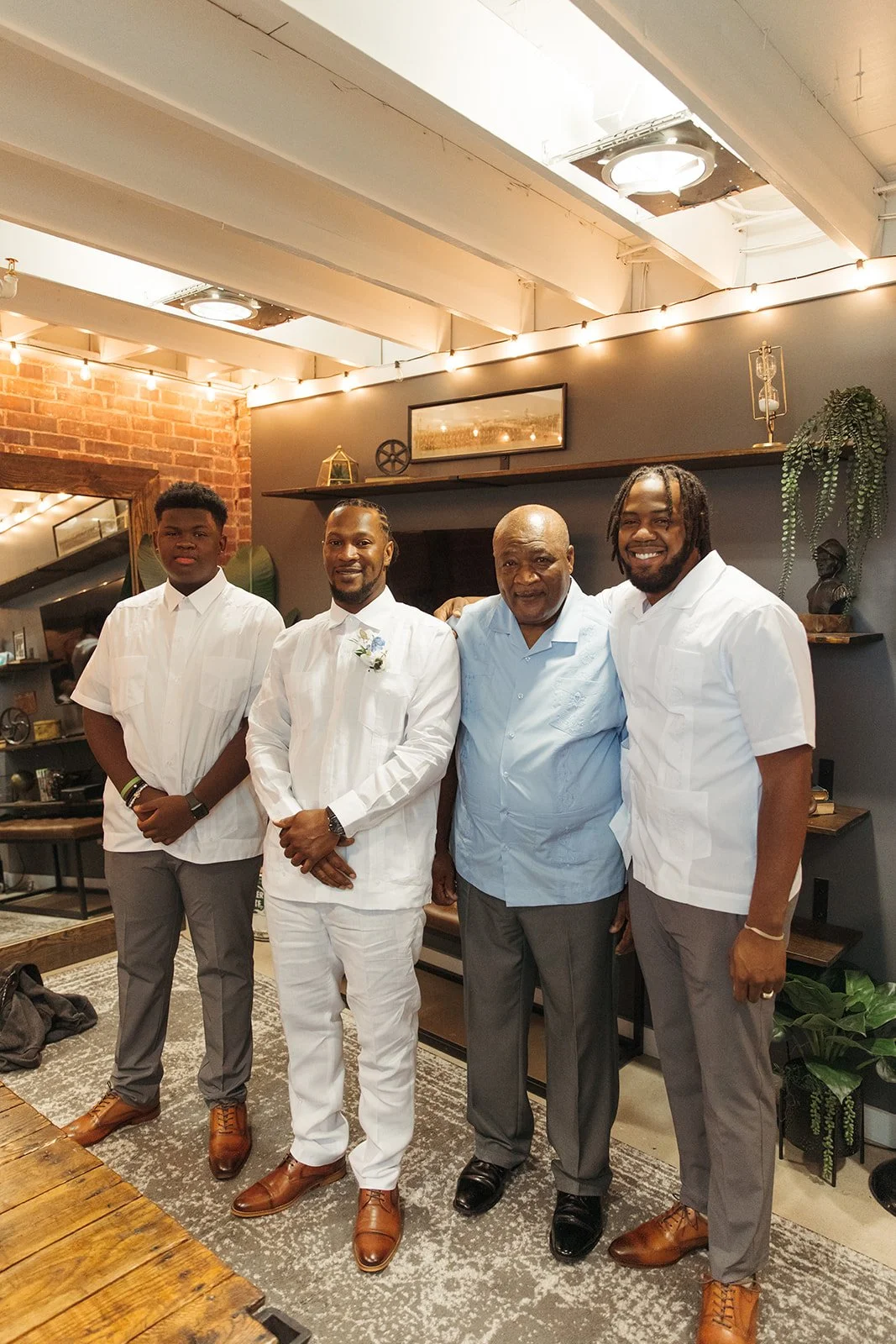 Four men standing together in a cozy, decorated interior space, dressed in formal and semi-formal clothing, smiling at the camera.