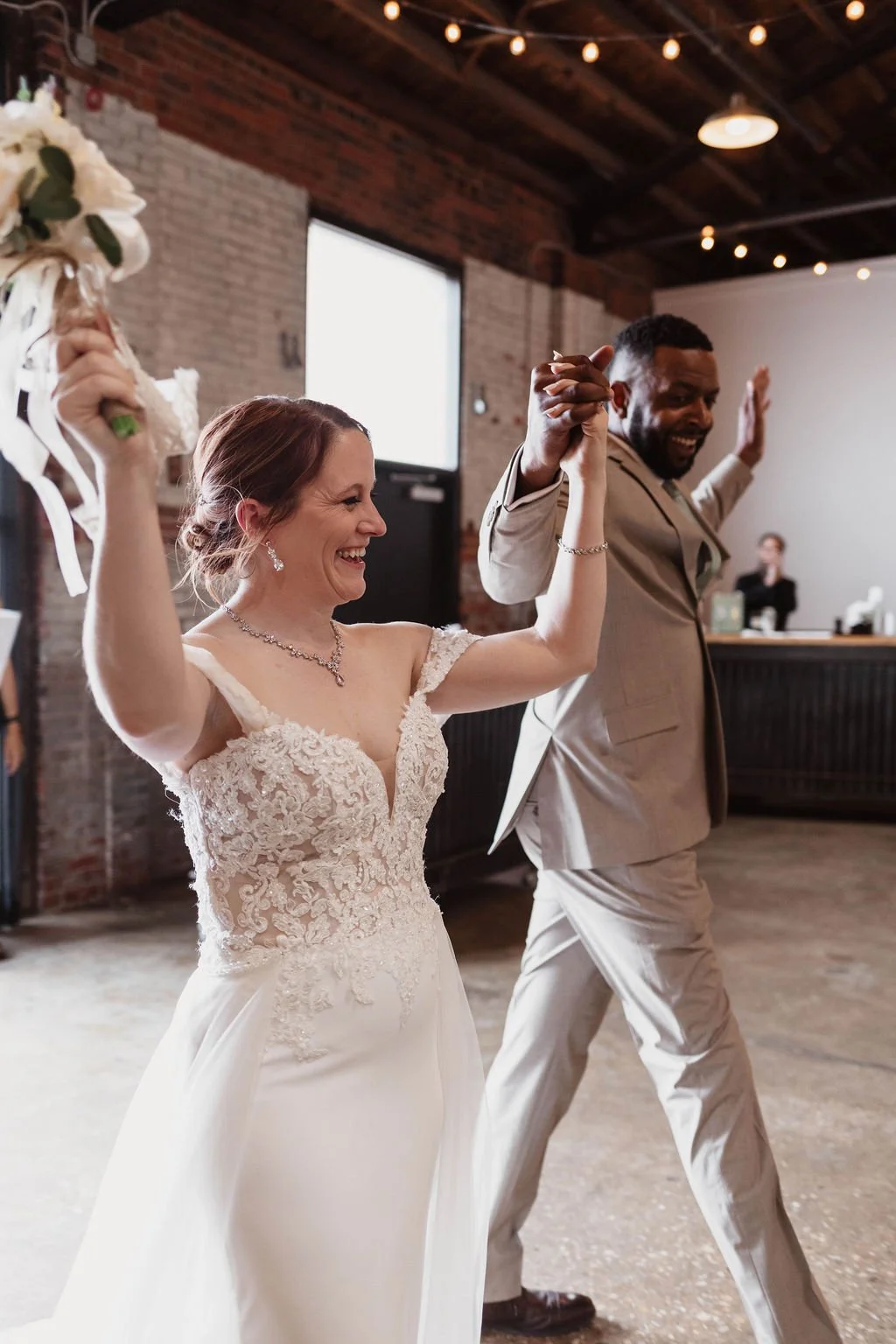 A bride and groom holding hands and celebrating at their wedding reception, smiling and dancing inside a rustic venue with exposed brick walls and string lights.