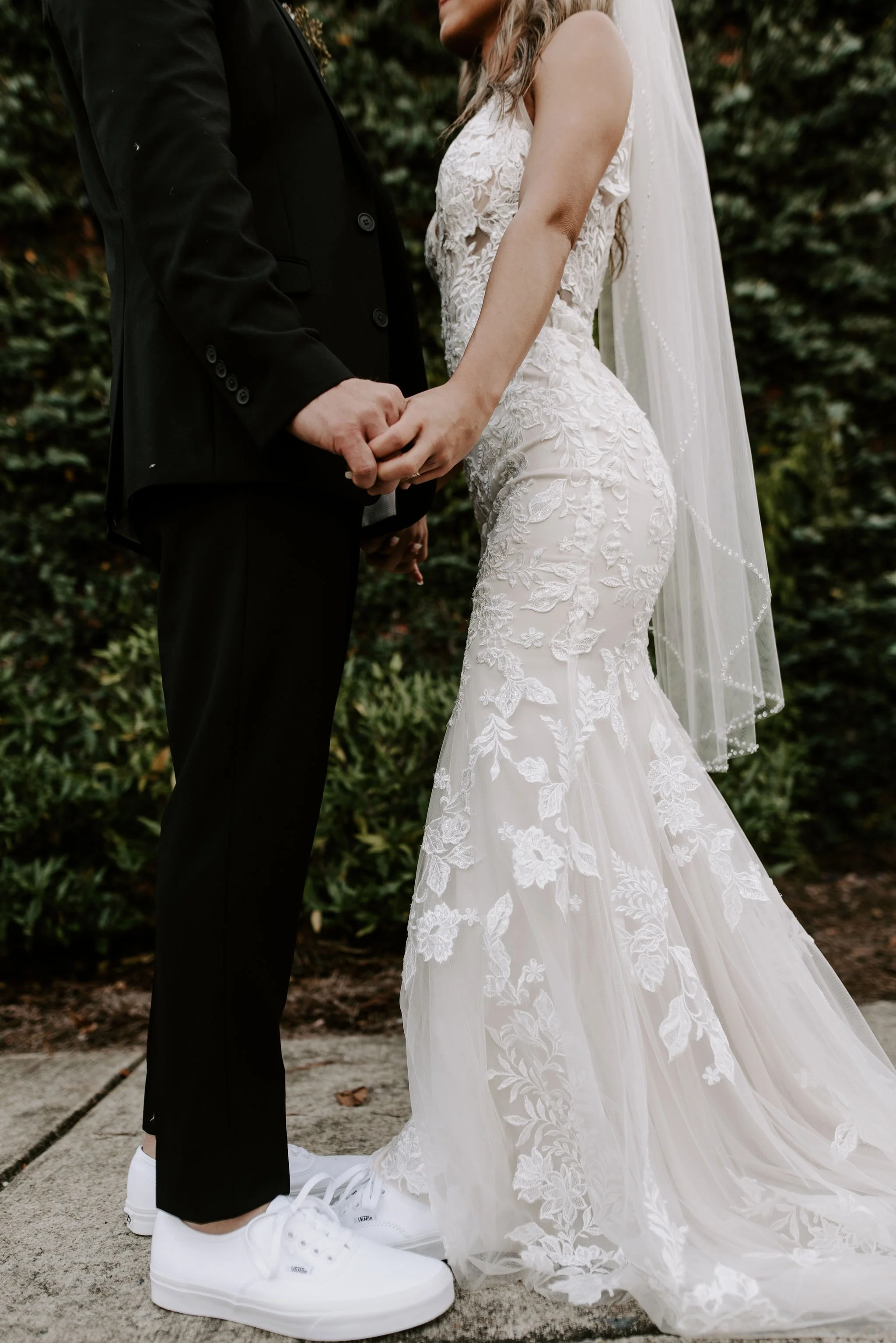 A bride and groom holding hands outdoors, with the bride in a lace wedding gown and veil, and the groom in a black suit and white sneakers.