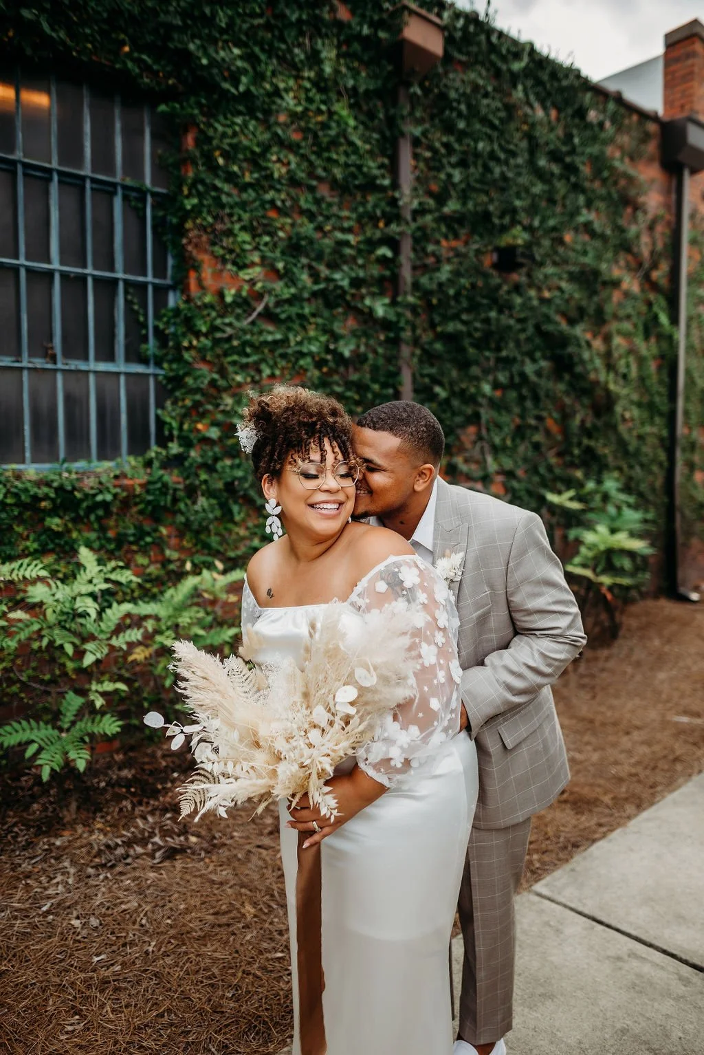 A joyful couple in wedding attire sharing a tender moment outdoors, with the woman holding a bouquet of dried flowers and the man embracing her from behind, against a background of green ivy-covered brick wall.