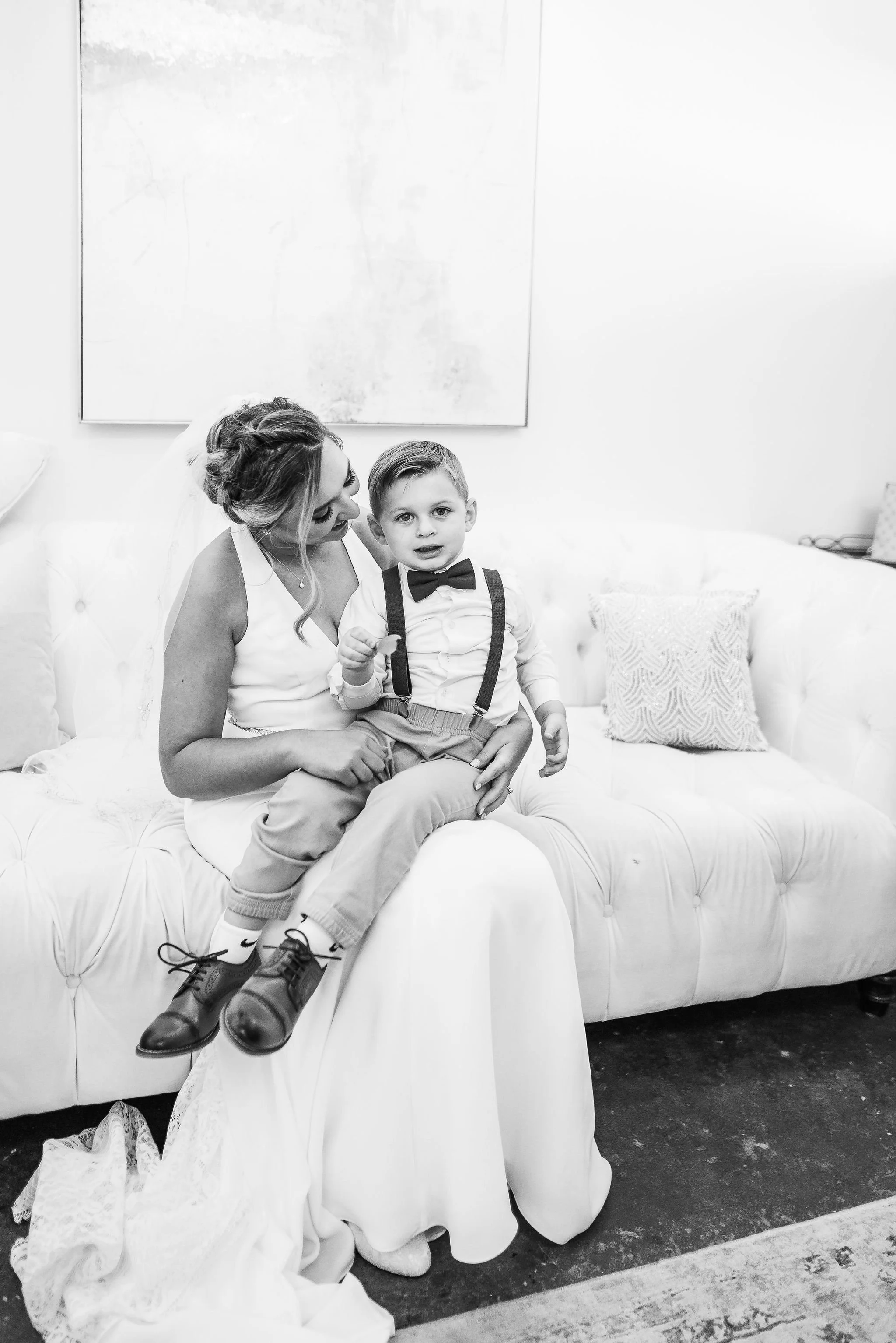 A woman, in a white dress, sitting on a tufted sofa, holding a young boy dressed in a white shirt, suspenders, bow tie, and beige pants, sitting on her lap. The woman is looking at the boy, who is looking at the camera in a black and white photo.