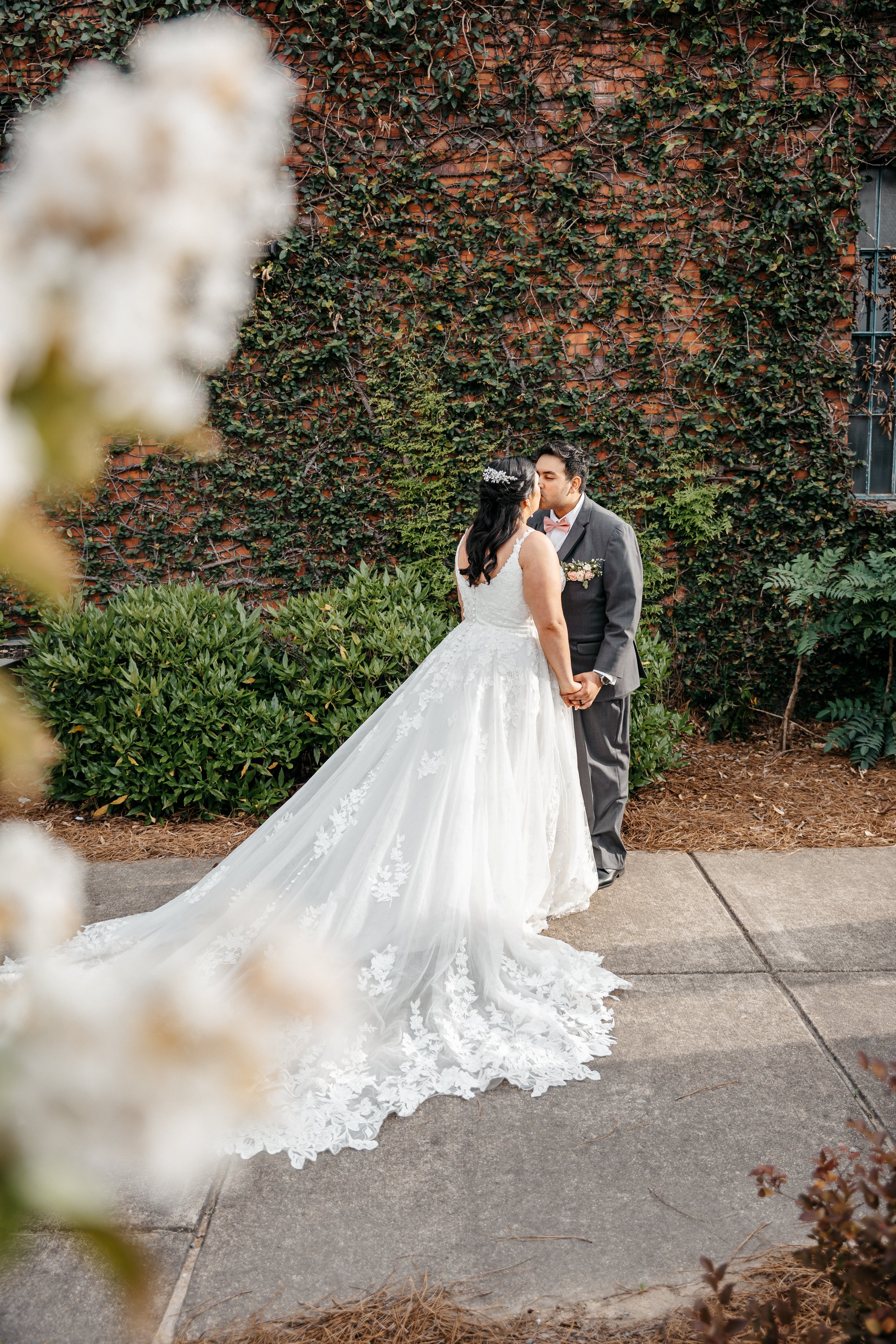 A bride and groom standing face to face, holding hands, about to kiss. The bride is wearing a white wedding gown with lace details, and the groom is dressed in a dark suit with a bowtie. They are outdoors with a brick wall covered in green ivy in the