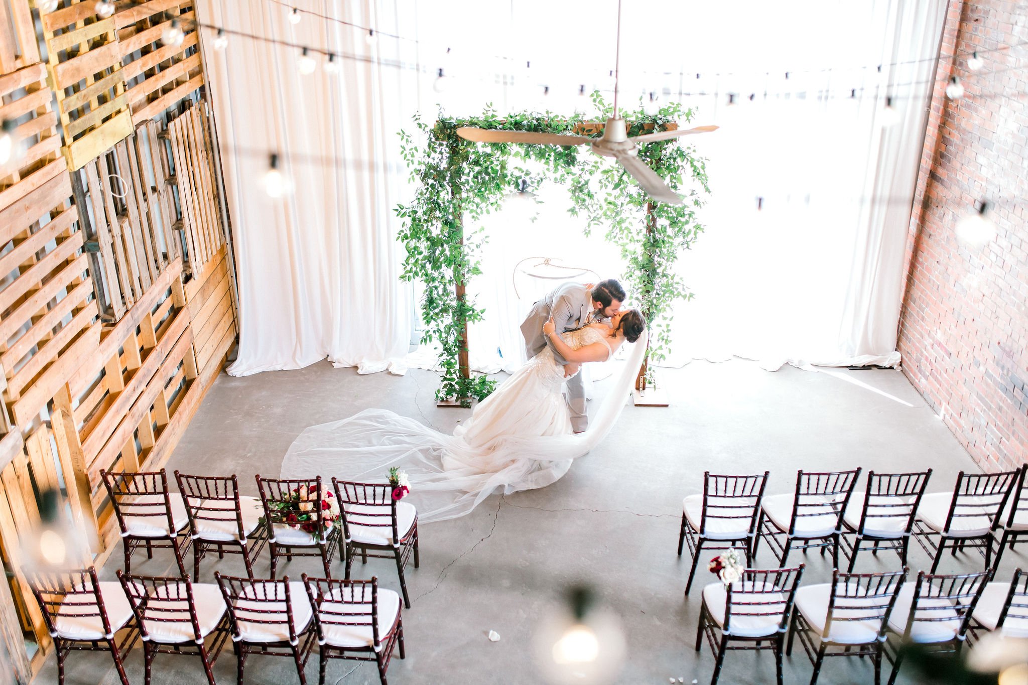A bride and groom at their wedding, sharing a kiss during the ceremony in a decorated indoor venue with wooden walls, string lights, and white curtains.