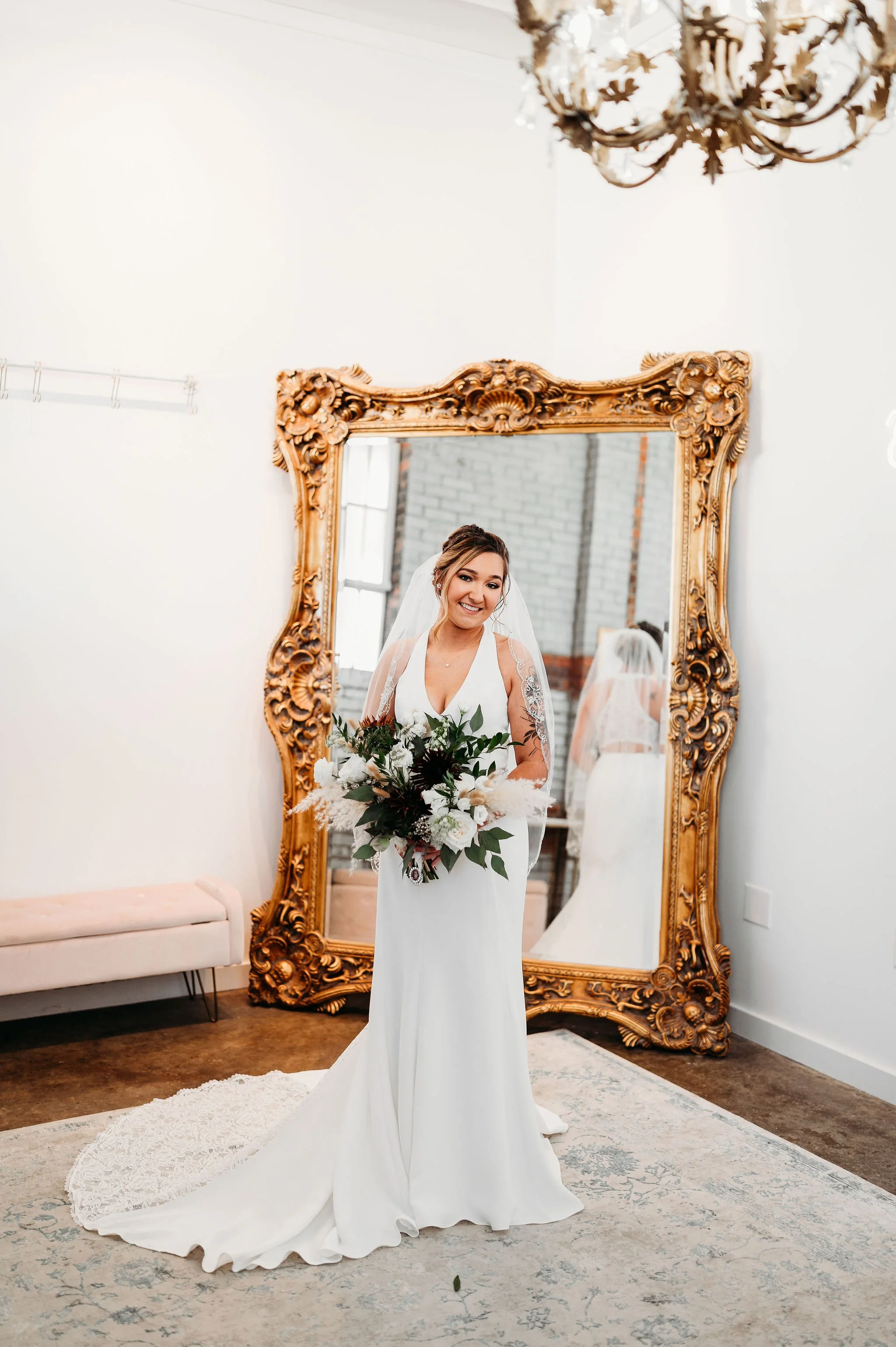 A bride in a white wedding dress smiling and holding a bouquet of flowers in a room with a large ornate mirror and a chandelier.