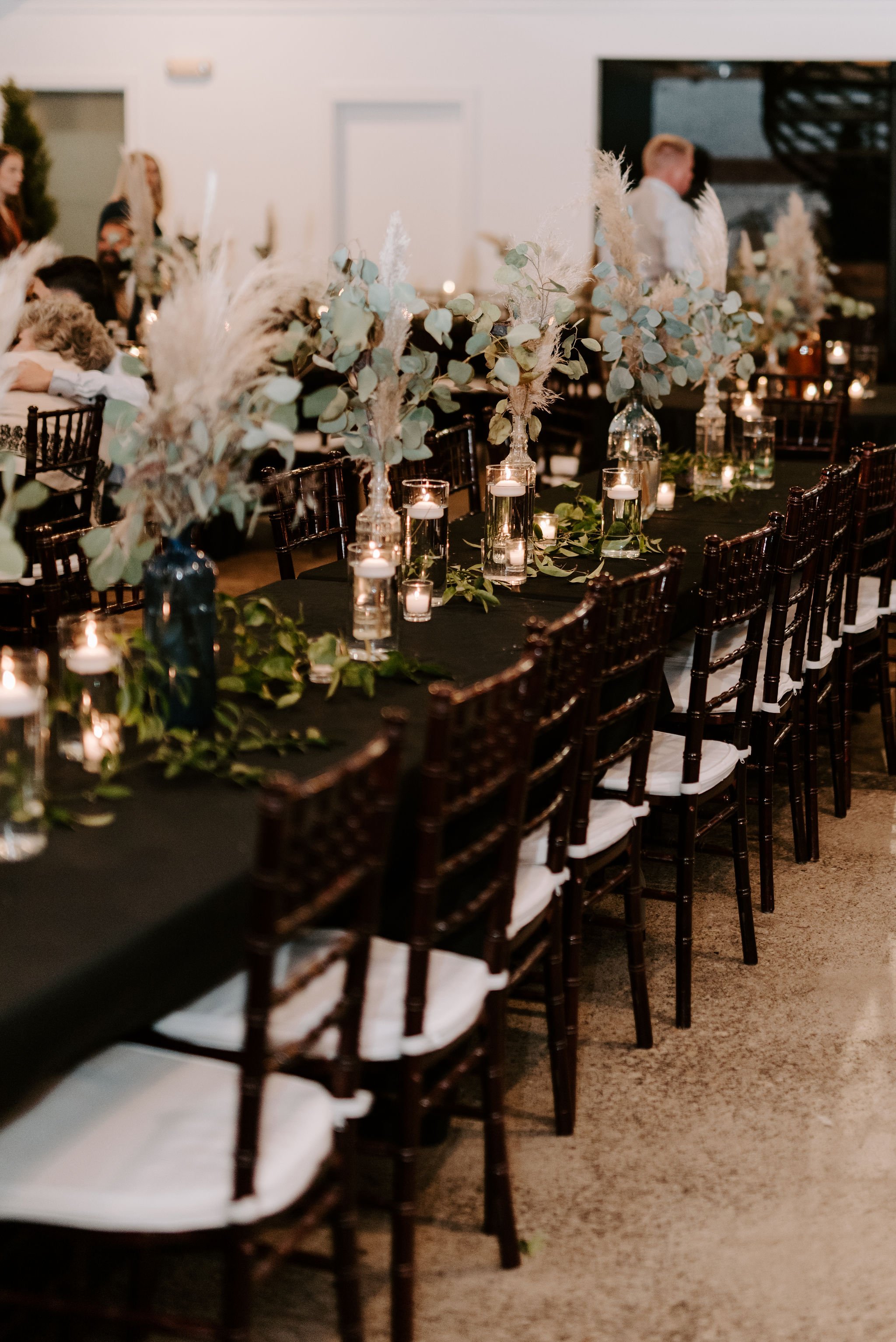 A decorated dining table at an indoor event with a black tablecloth, candle centerpieces in glass holders, and tall floral arrangements with eucalyptus and pampas grass in dark vases.