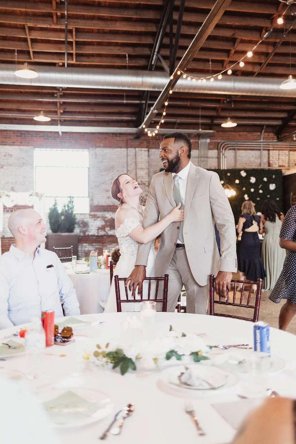 A wedding reception with a bride and groom sharing a joyful moment with a guest standing and laughing, in a rustic venue with exposed brick walls and string lights.