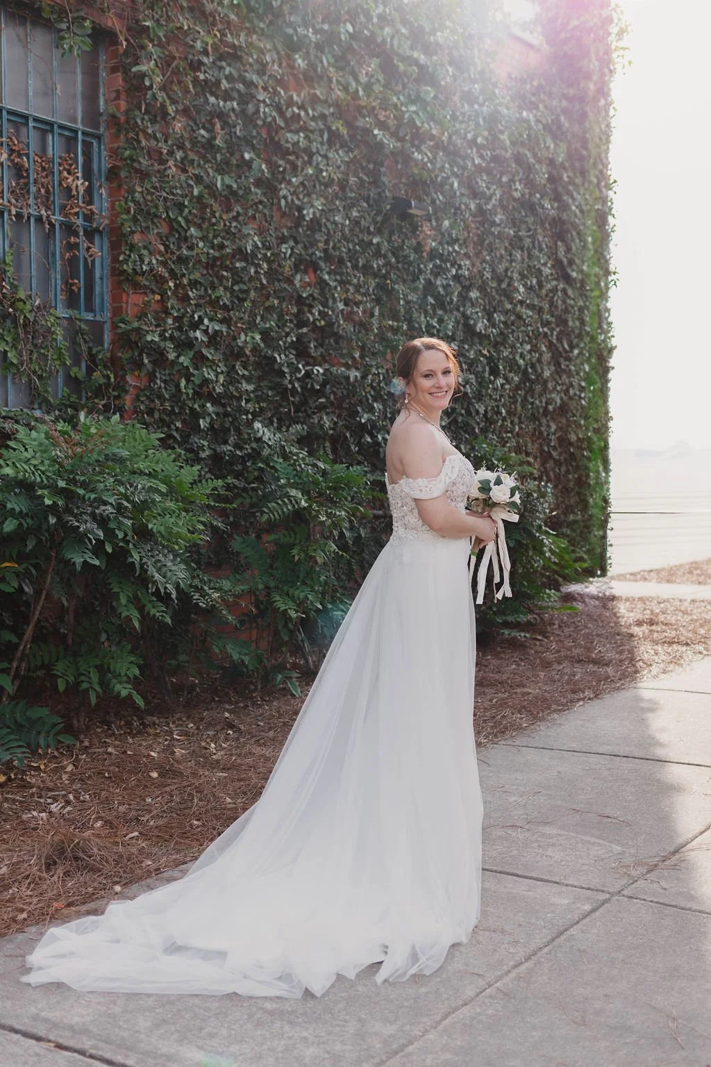 A bride in a white wedding dress holding a bouquet, standing next to a brick wall covered with ivy, smiling at the camera with sunlight shining in the background.
