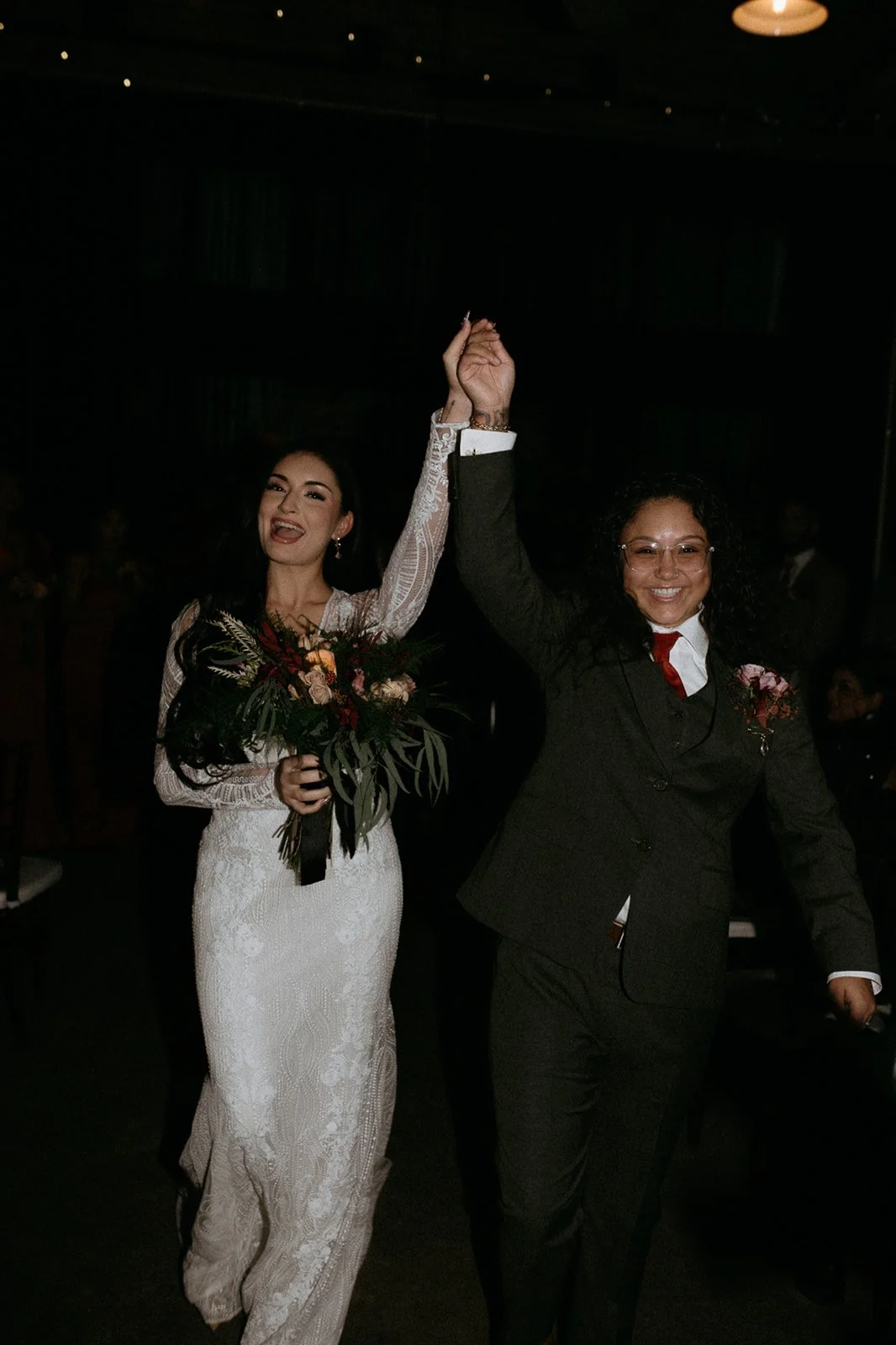 Two women, one in a white wedding dress holding a bouquet, and the other in a dark suit, are smiling and holding hands up in celebration at a wedding reception.