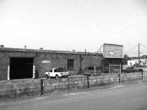 A black-and-white photo of a small commercial building with a parking lot in front, cars parked along the street, and power lines in the background.