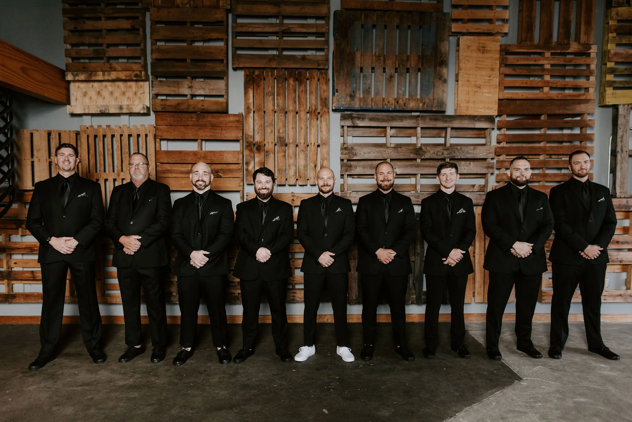 A group of nine men in black suits standing in a line, smiling, with a wooden pallet wall background.
