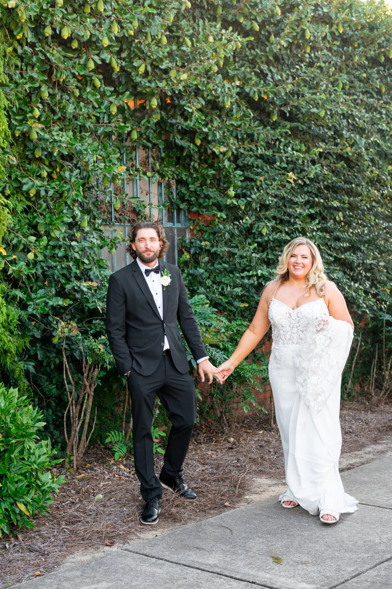 A couple dressed in wedding attire standing outdoors on a sidewalk, holding hands, with a lush green background.