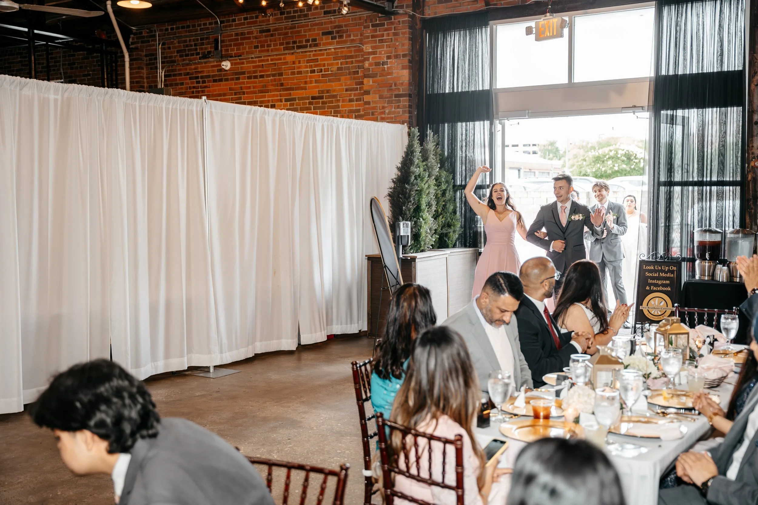 People at a wedding reception, some seated at a banquet table clapping, a woman in a pink dress and two men in suits are standing and smiling near a white curtain backdrop, with two women and two men in formal attire behind them, in a venue with bric