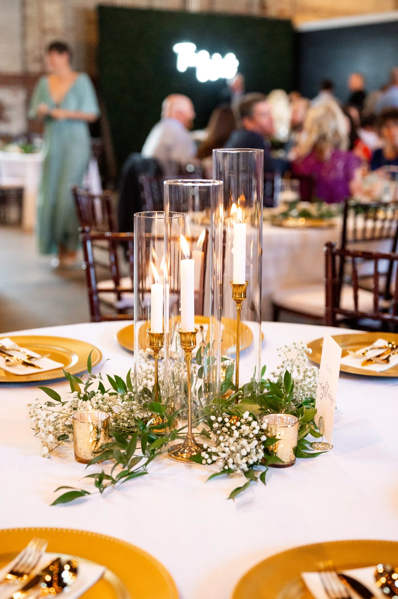 A wedding reception table with gold and white decor, tall glass candle holders with white candles, small gold votive holders, green foliage, and white baby's breath flowers. In the background, blurred guests are seated, and a neon sign on a dark wall