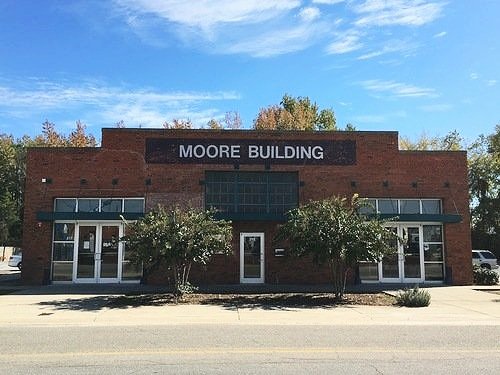 A brick building with a sign that reads 'Moore Building' on the front, two trees in front, and glass doors and windows.