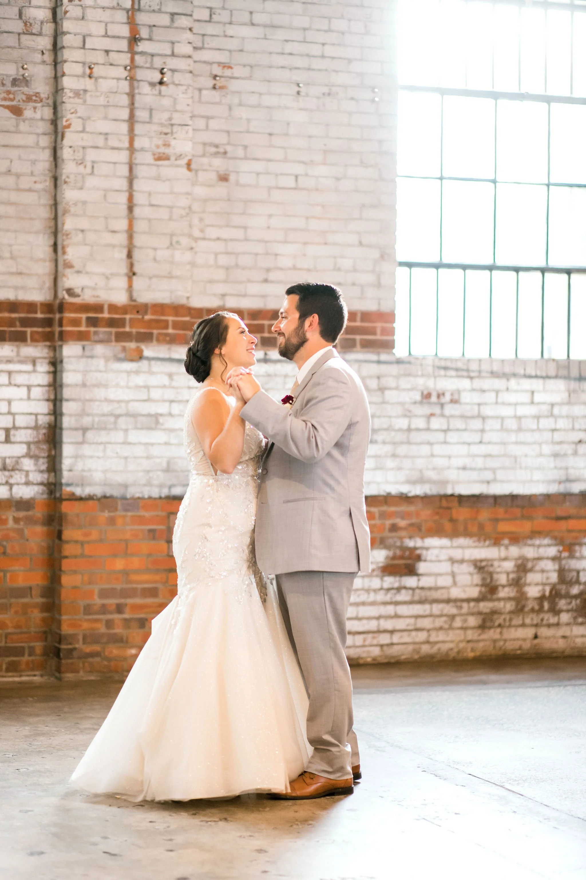 A bride and groom dancing together in an industrial-style building with large windows and brick walls.