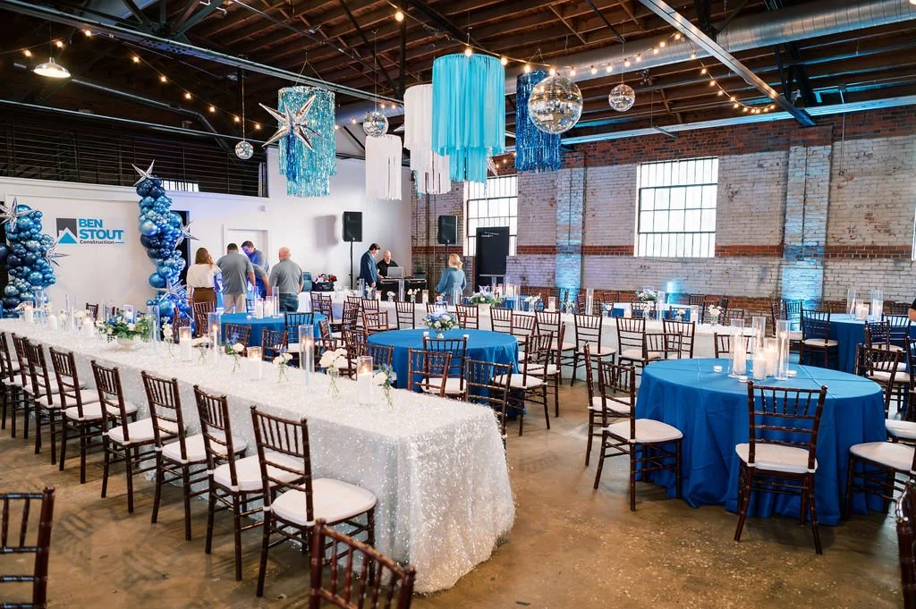 Decorated event space with blue, white, and silver decorations, round and rectangular tables with blue and white tablecloths, balloons, hanging streamers, disco balls, and candles, with a few people preparing the setup.