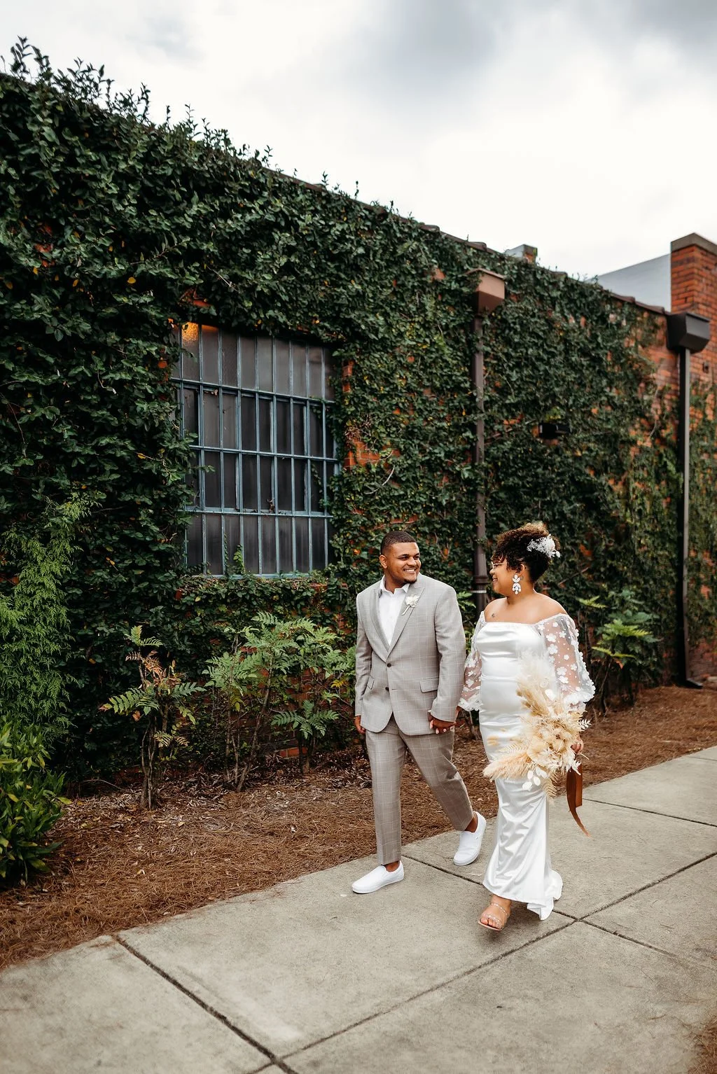 A bride and groom holding hands and walking outside near a brick building covered in greenery, smiling at each other on their wedding day.