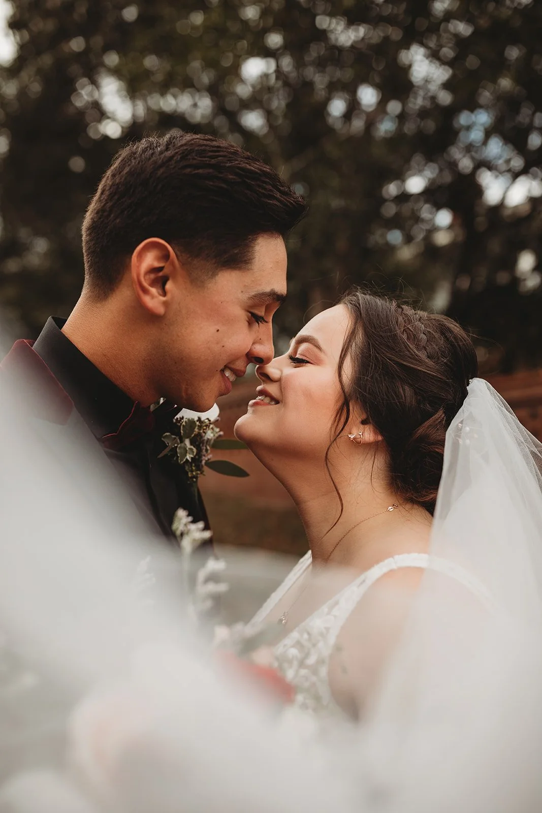 Close-up of a bride and groom about to kiss, smiling, with trees in the background.