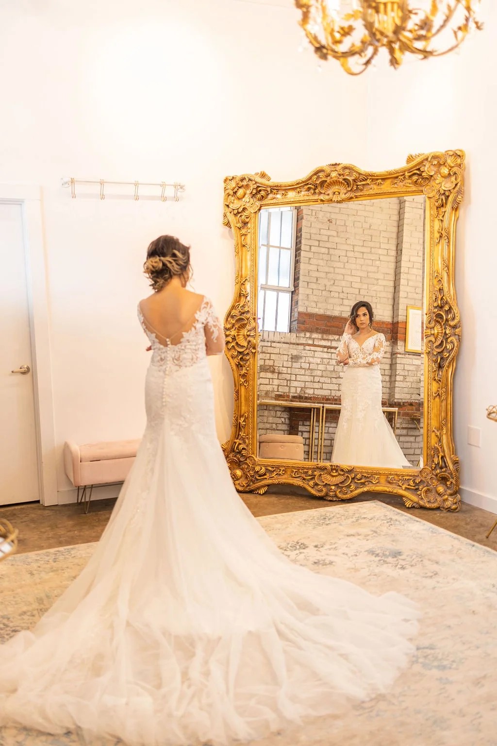 Bride in a wedding dress looking in a large ornate gold mirror, with her reflection visible.