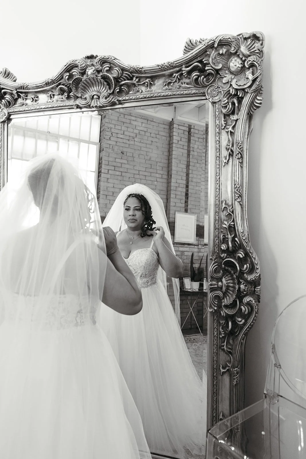 A bride in a wedding gown and veil is looking in a mirror, adjusting her hair.