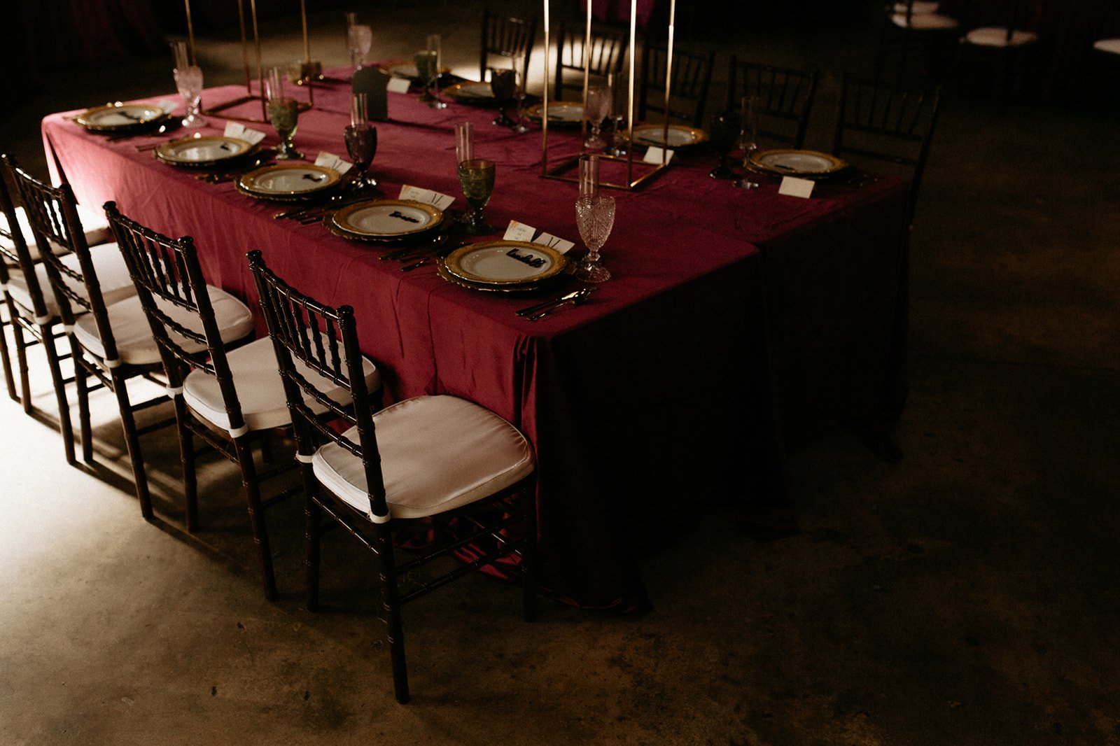 A large dining table set for a formal event with a red tablecloth, gold-trimmed plates, crystal glasses, black and gold utensils, and white cushioned chairs, in a dimly lit room.