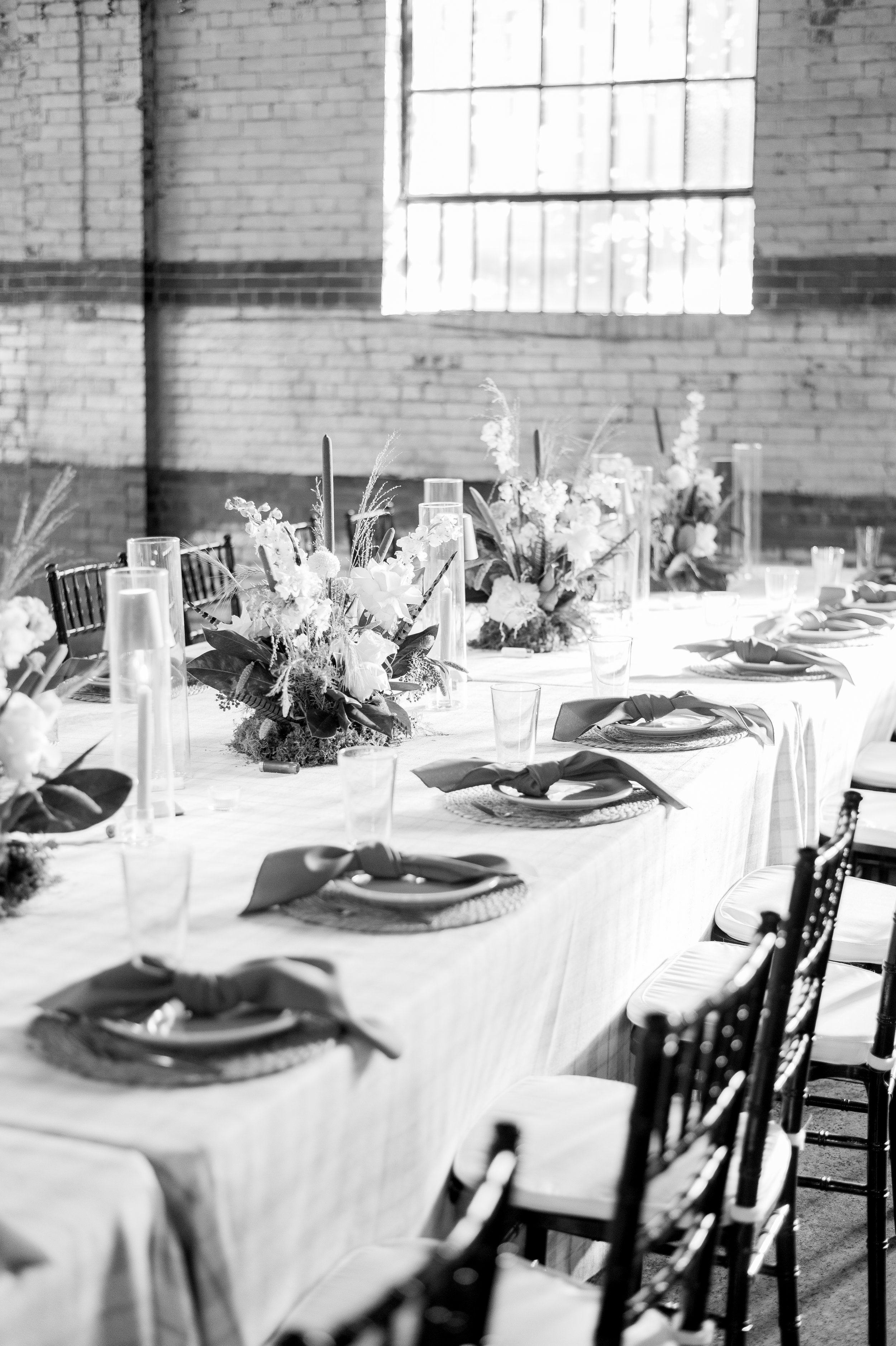 A long banquet table set with floral centerpieces, plates with napkins, drinking glasses, and tall candles, in an industrial-style room with brick walls and large windows, in black and white.