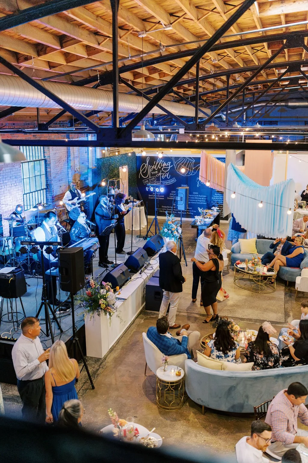 Indoor celebration with live band, people dancing, and socializing under warm string lights, with decorated tables and seating areas.