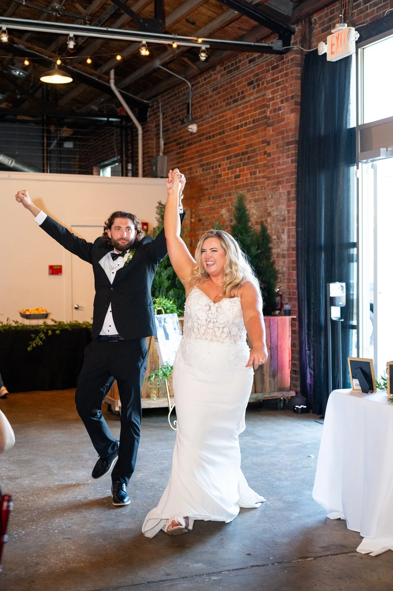 A bride and groom dancing at their wedding reception in a rustic venue with exposed brick walls, black curtains, and greenery, celebrating joyfully.