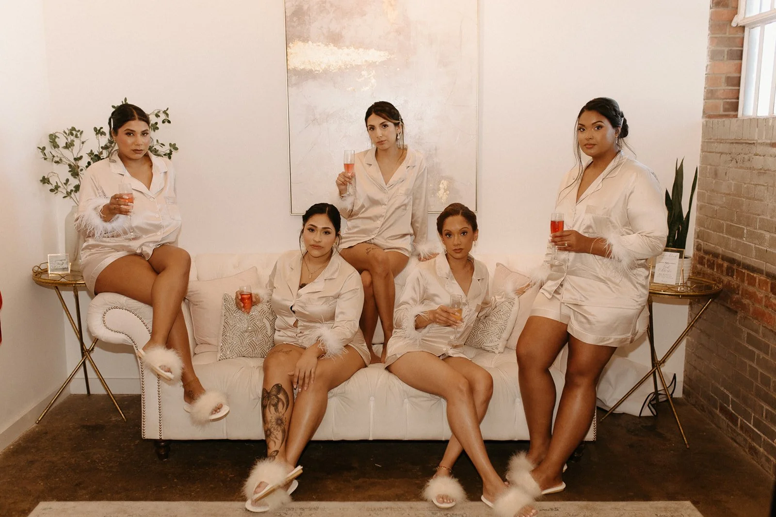 Six women in matching satin pajamas with feather trim, wearing fluffy slippers, sitting and standing on and around a cream tufted sofa in a living room, holding glasses of bubbly, with a modern art piece on the wall behind them.