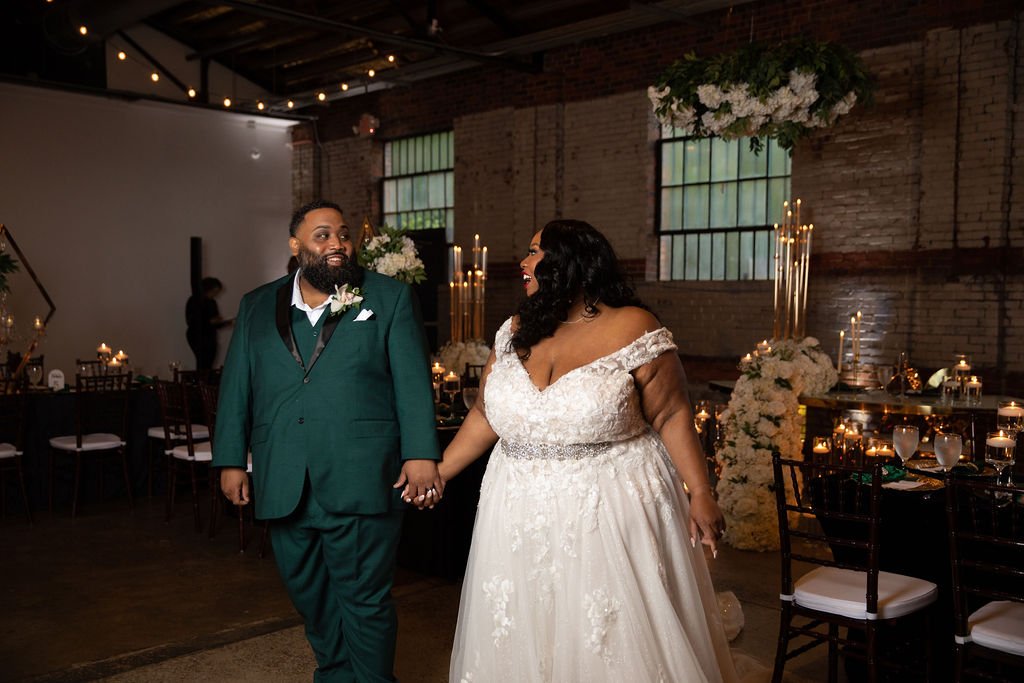 A bride and groom holding hands and smiling at each other in a decorated wedding reception venue with candlelit tables and floral arrangements.