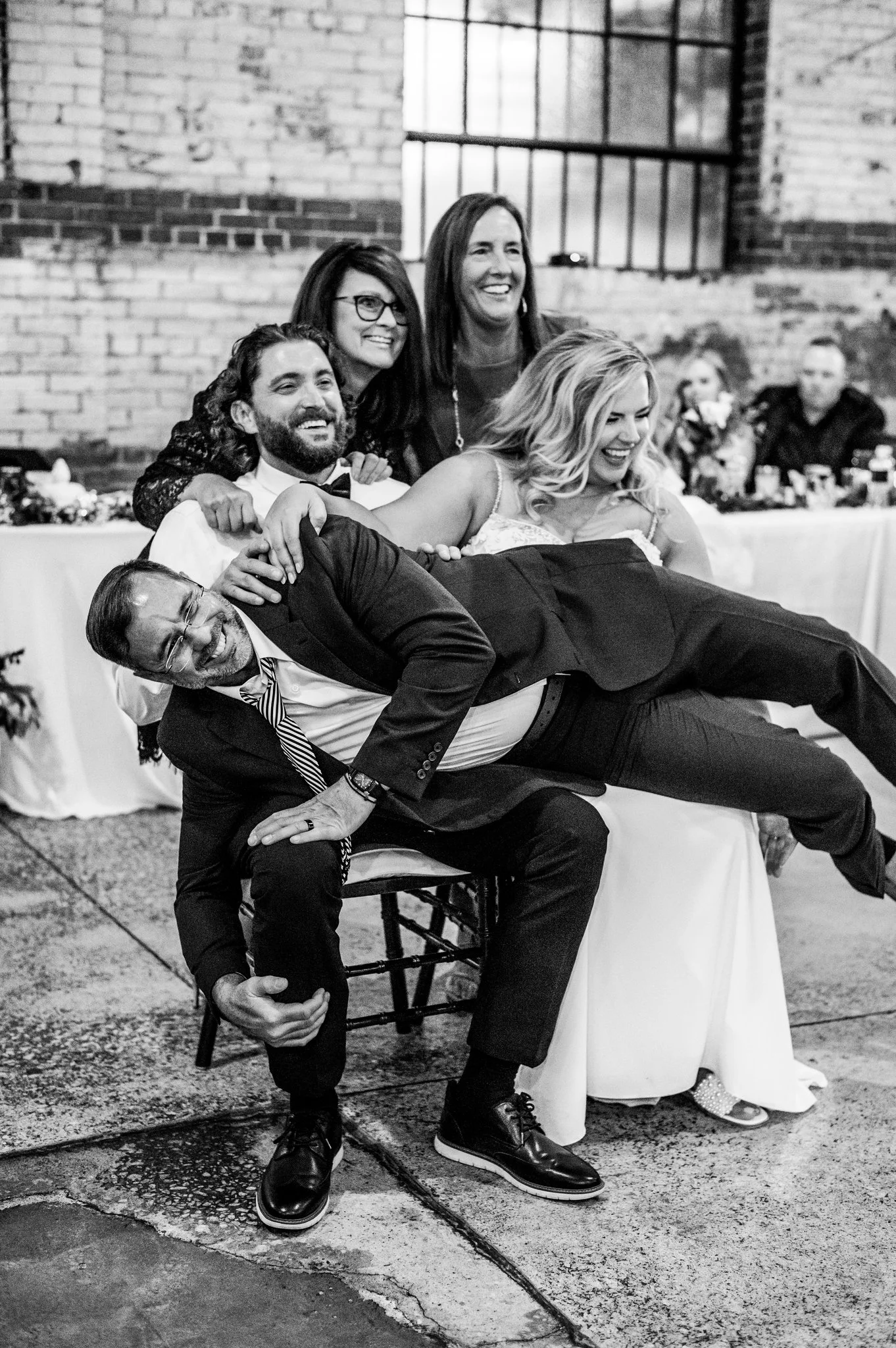 Group of five people, including a bride and groom, posing playfully at a wedding reception in a rustic indoor venue with exposed brick walls. The groom is lying on the lap of a seated man, with others smiling around them.