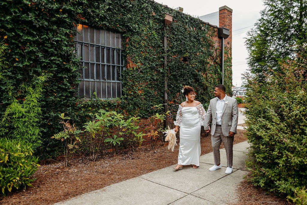 A couple dressed in wedding attire walking hand in hand on a sidewalk surrounded by green bushes and ivy-covered brick building.