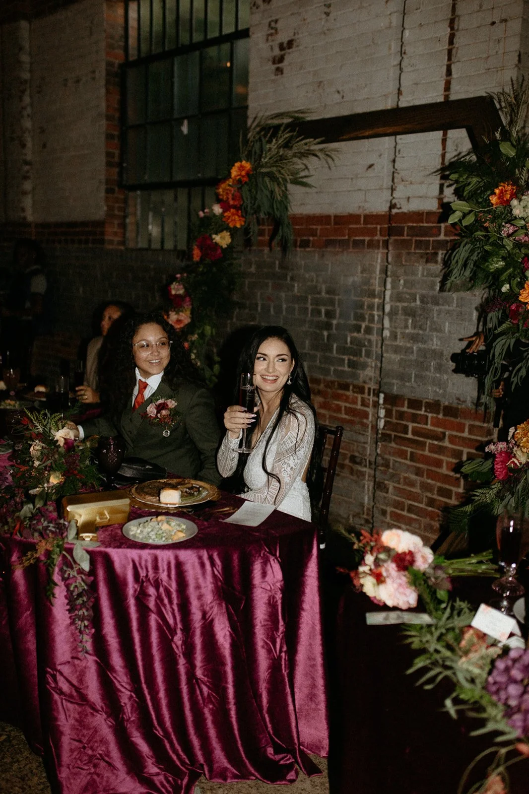 Two women sitting at a decorated table with flowers, one holding a drink, at a celebration or event in a brick-walled venue.