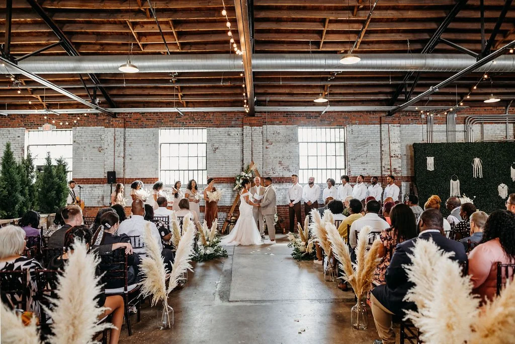Wedding ceremony with bride and groom standing in front of officiant, guests seated on both sides, inside a rustic industrial venue with exposed brick walls and large windows, decorated with pampas grass arrangements.