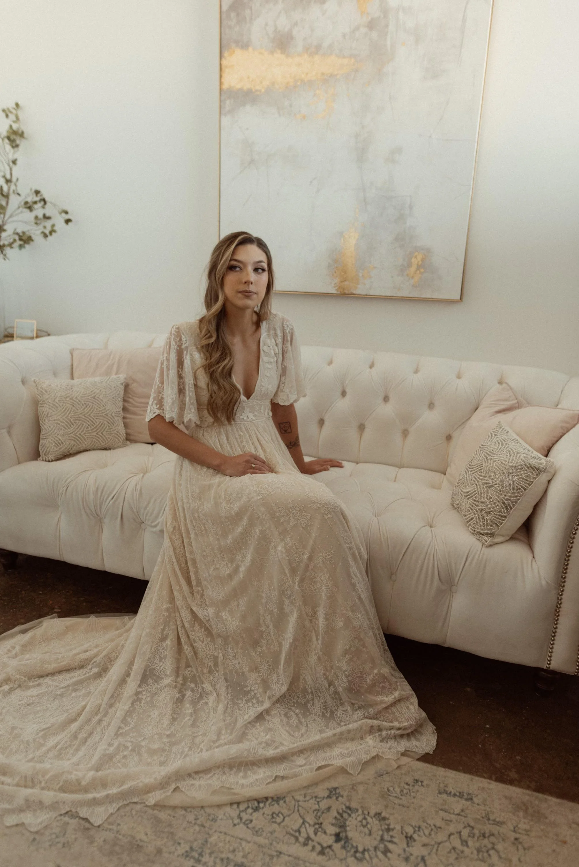 A woman in a cream-colored lace wedding dress sitting on a cream tufted sofa in a modern, minimalist living room with abstract wall art and decorative pillows.
