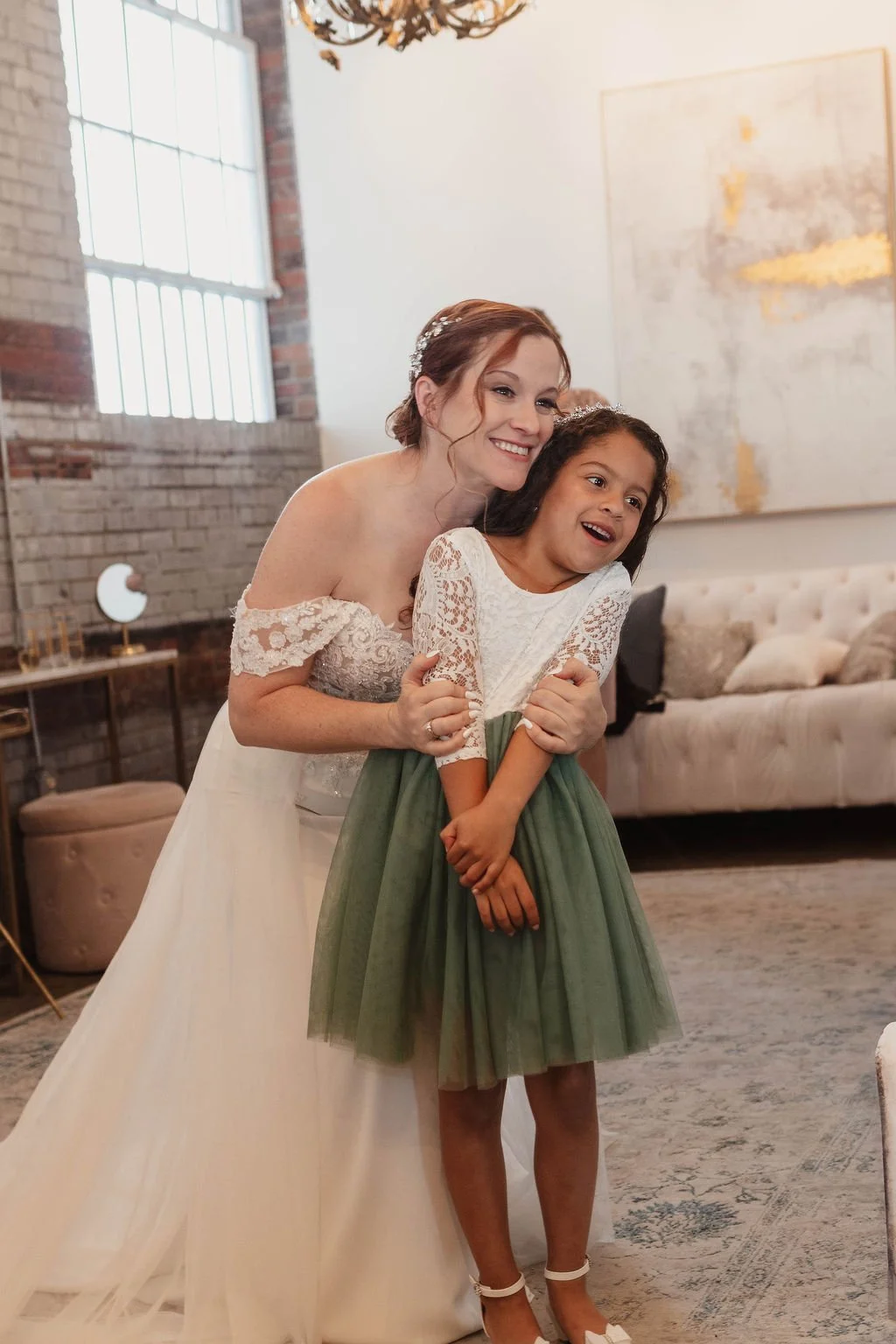 A woman in a wedding dress hugging a young girl in a white lace top and green skirt in a warmly decorated room with a sofa, large window, and abstract art on the wall at Studio 215 a modern warehouse venue in Fayetteville, NC