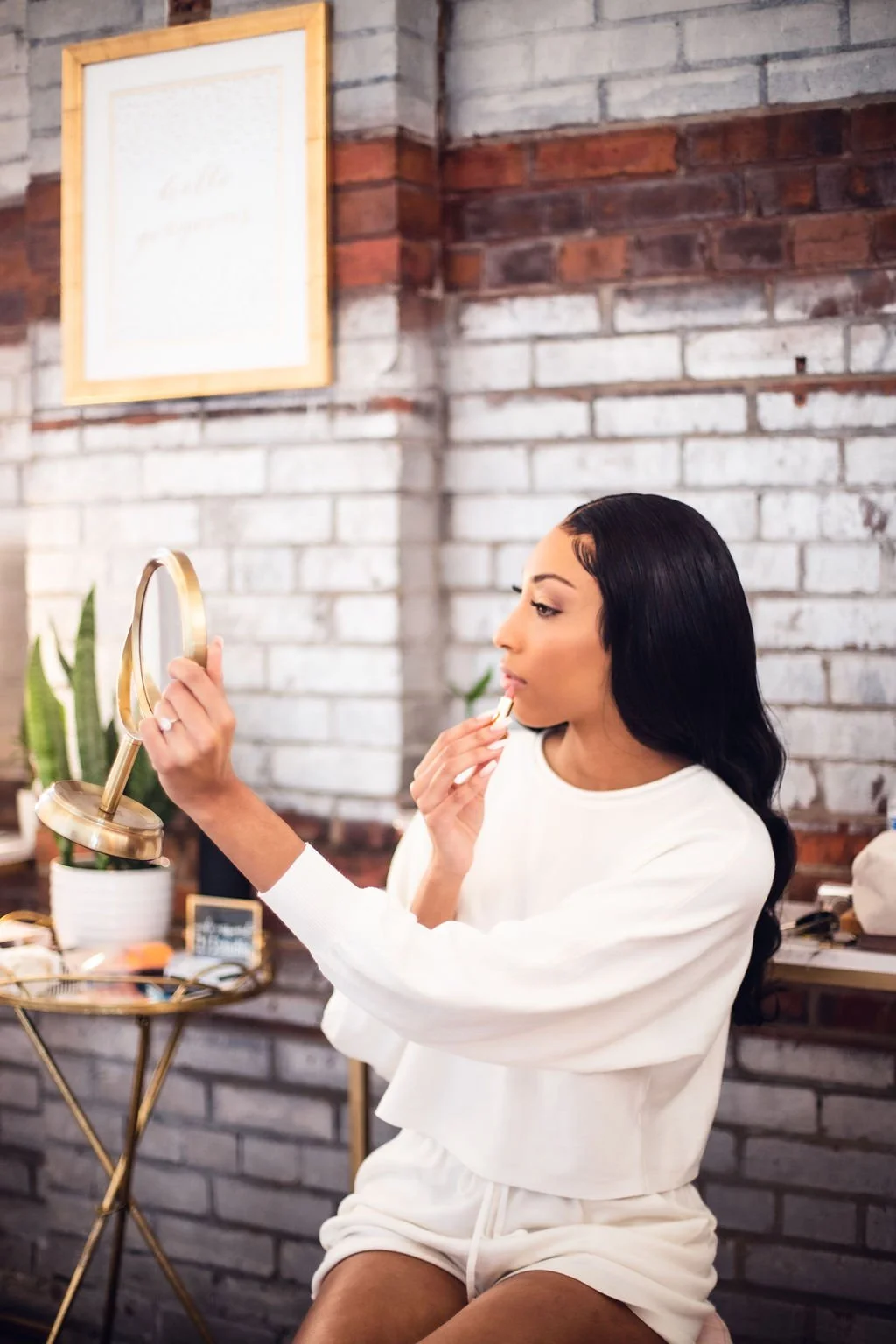 Woman applying makeup while looking into a mirror in a cozy, modern room with brick walls and indoor plants.