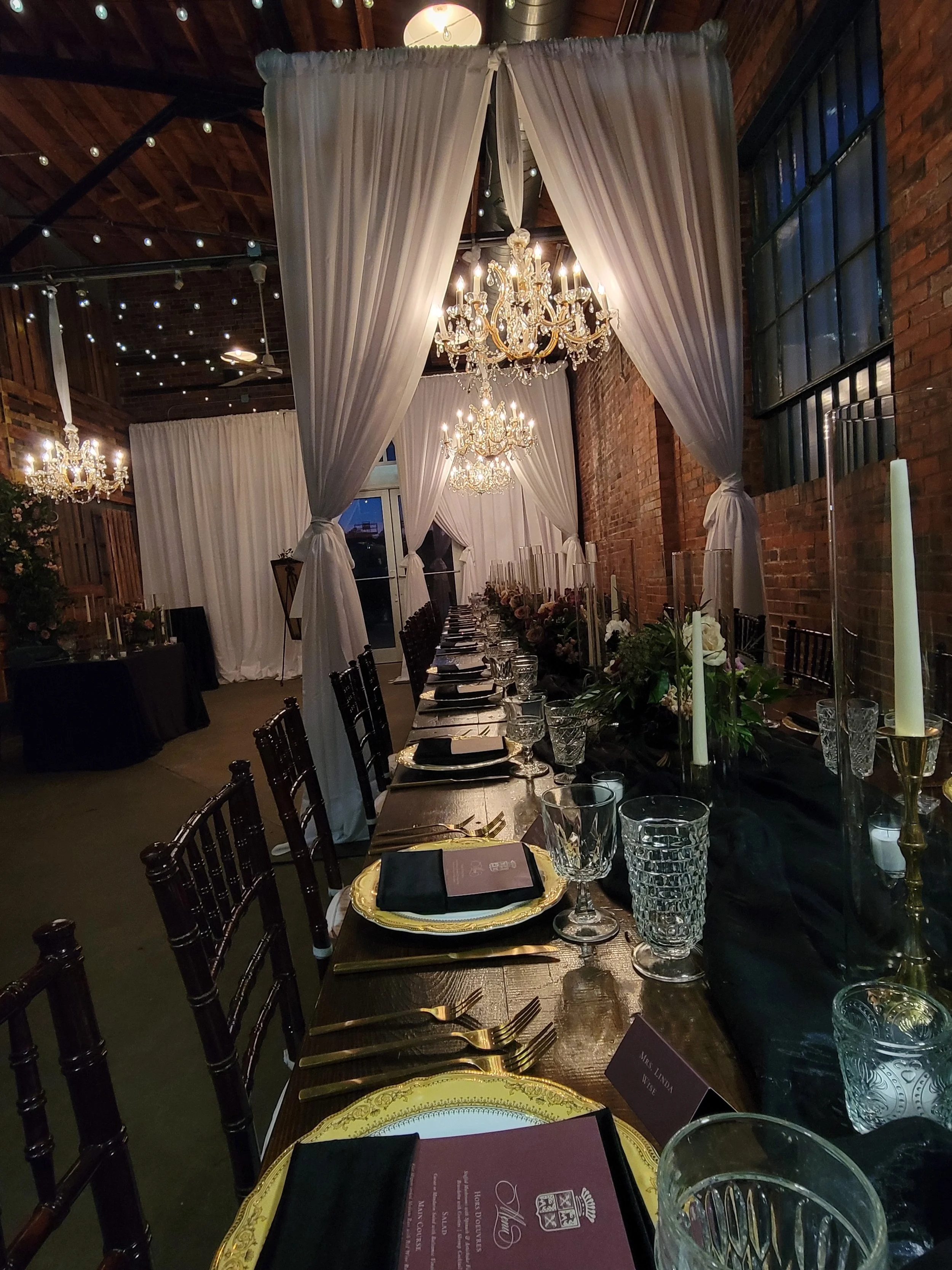 An elegantly decorated dining table set for a formal event, featuring black chairs, gold-rimmed plates, crystal glasses, black napkins, and tall white candles in gold candleholders. The table is adorned with a floral centerpiece, and the background includes white drapes, chandeliers, string lights, and brick walls, creating a luxurious atmosphere.