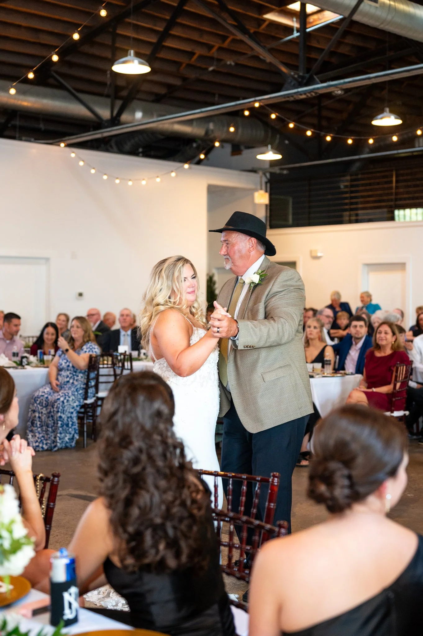 A bride and an older man are dancing at a wedding reception. The man is wearing a hat and a tan suit with a white boutonniere. The bride has long blonde hair and is wearing a white wedding dress. There are guests seated at tables in the background, a