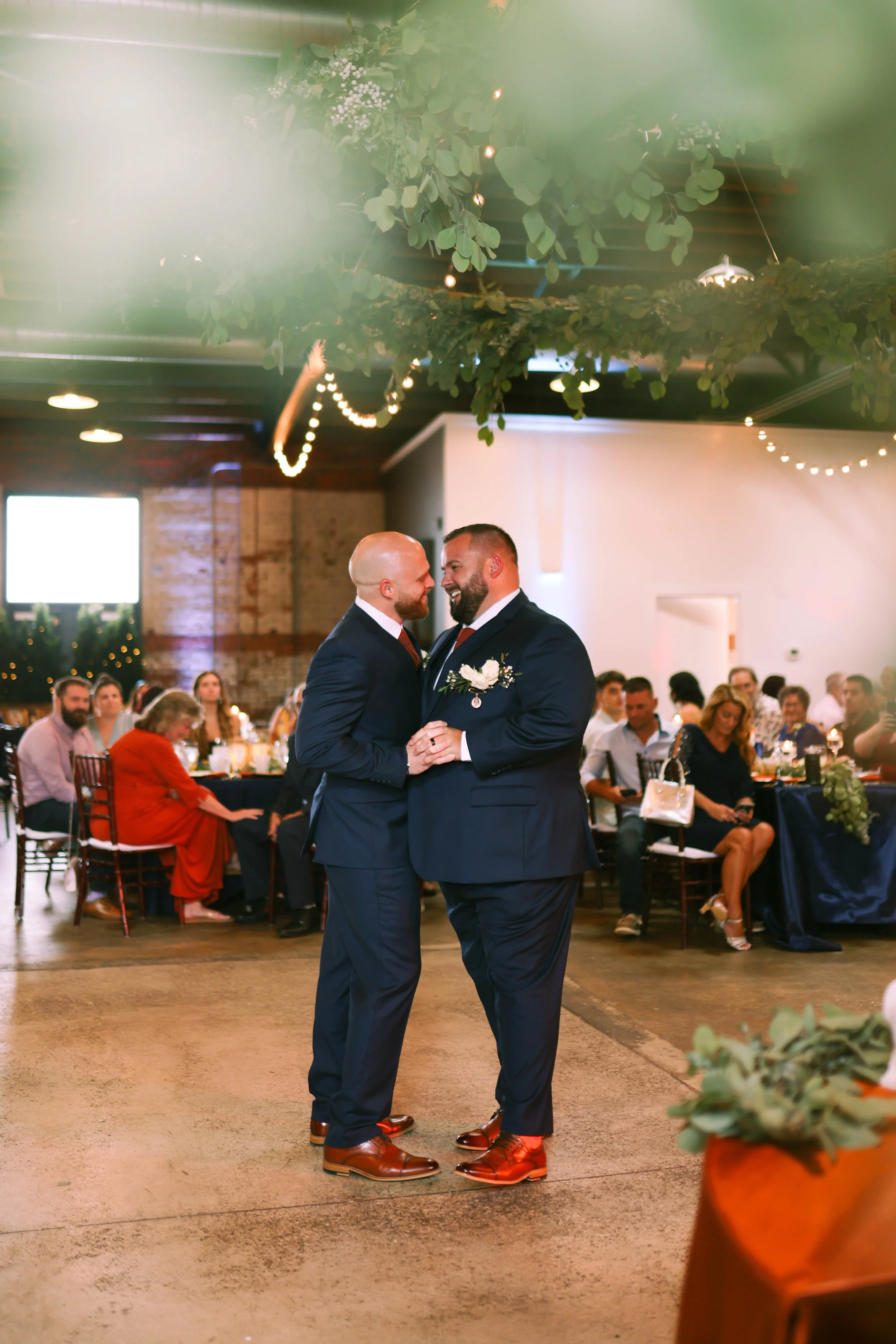 Two men in suits, one with a bald head and beard, the other with dark hair and beard, smiling and holding hands, sharing a dance at a wedding reception. Guests seated at tables are visible in the background.