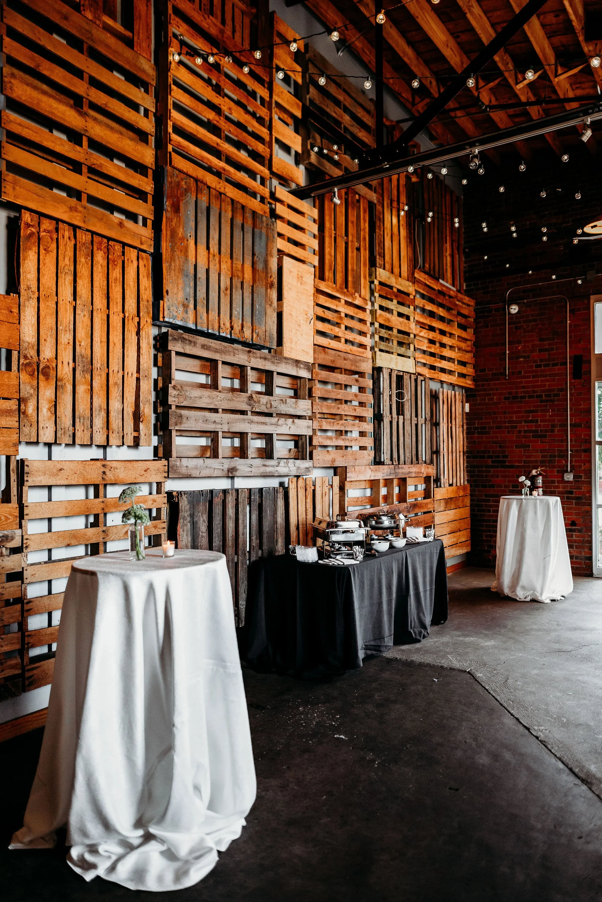 Interior of a rustic event space featuring a wall decorated with wooden pallets of various sizes, a buffet table with chafing dishes, and two tall cocktail tables with white tablecloths, small candle, and vase with greenery.