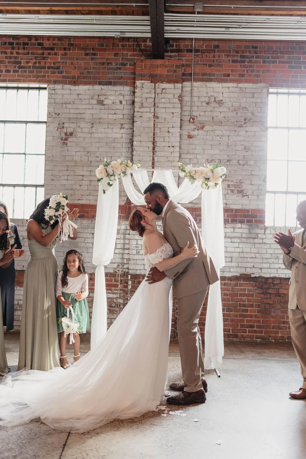 A couple kissing during their wedding ceremony in front of a floral decorated arch, with guests clapping and smiling. The bride is wearing a white wedding gown, and the groom is in a beige suit. The ceremony is held in a rustic venue with exposed bri
