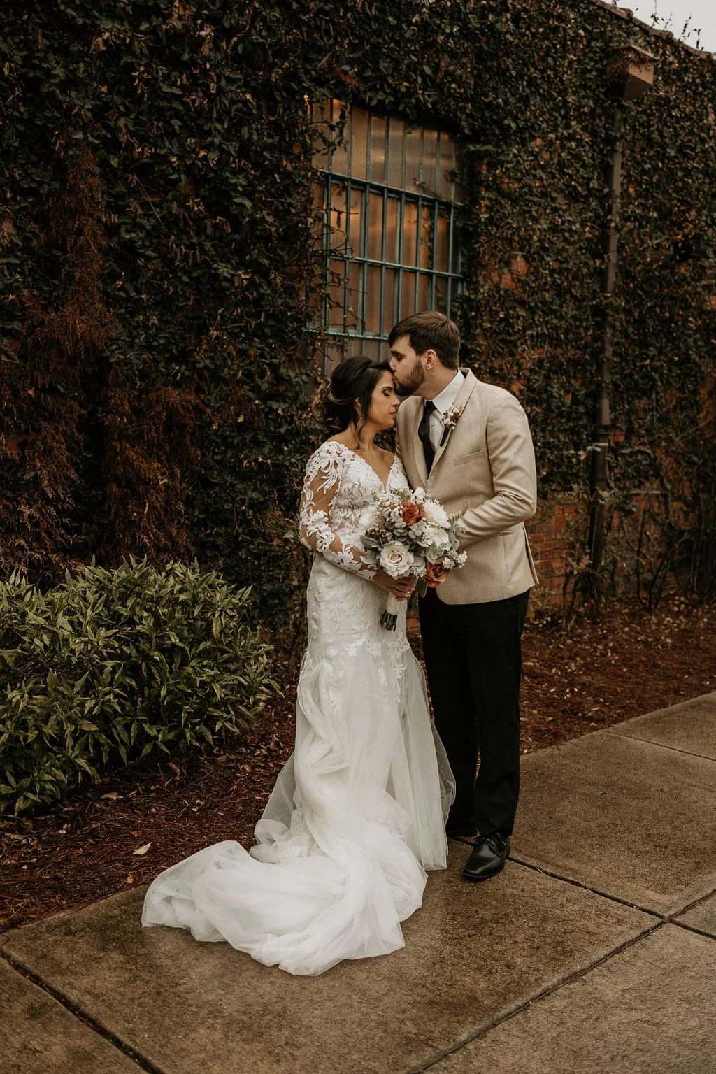 Bride and groom posing in front of the signature green ivy wall at Studio 215 in downtown Fayetteville, NC; a lush urban garden photography backdrop at our industrial warehouse venue near Fort Bragg and Raleigh.