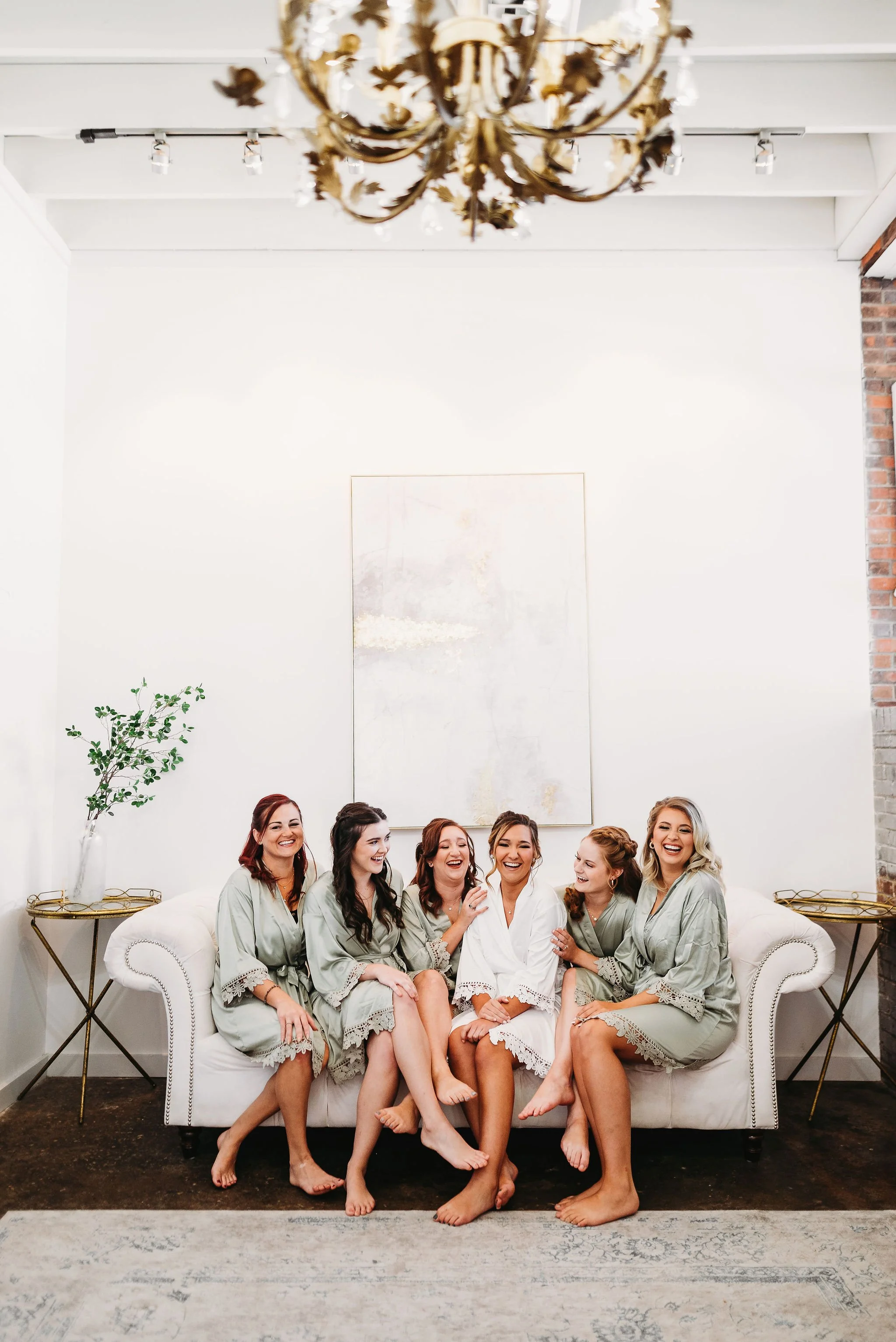 Group of six women in robes sitting on a white couch, smiling, in a bright, modern living room with a chandelier, artwork on the wall, and side tables with decorative items.