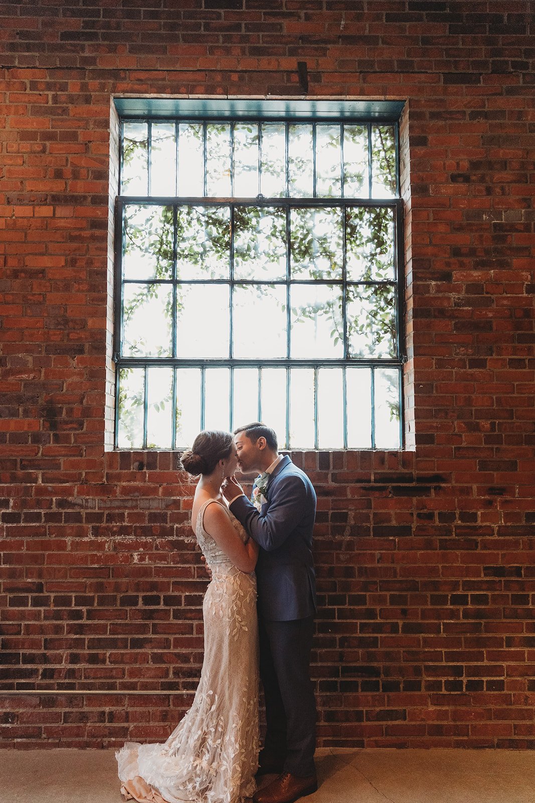 A bride and groom standing close, sharing a kiss inside a brick building with a large window above them showing trees outside.