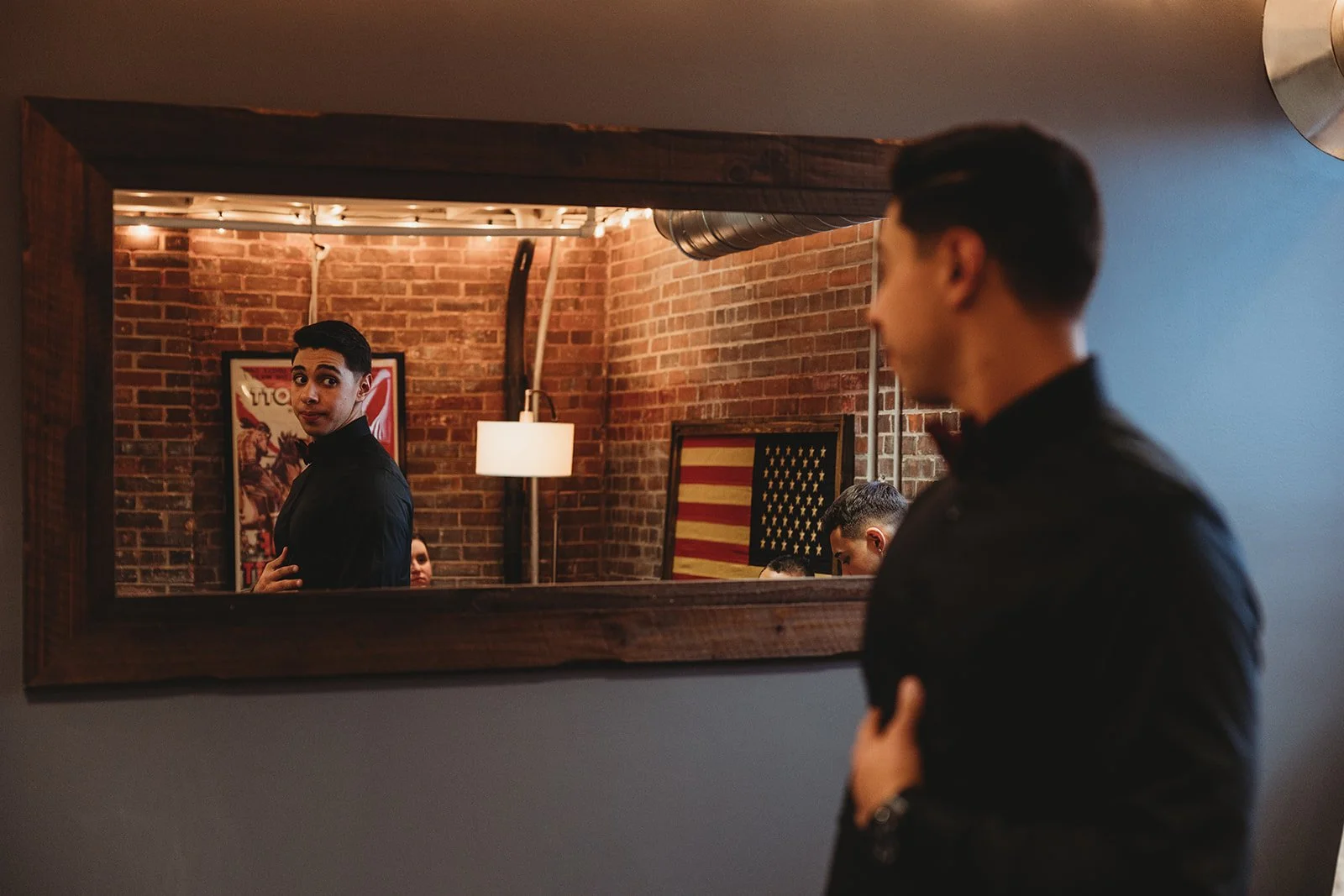 A man looking at his reflection in a wall mirror, with a brick wall and American flag art behind him, inside a dimly lit room.