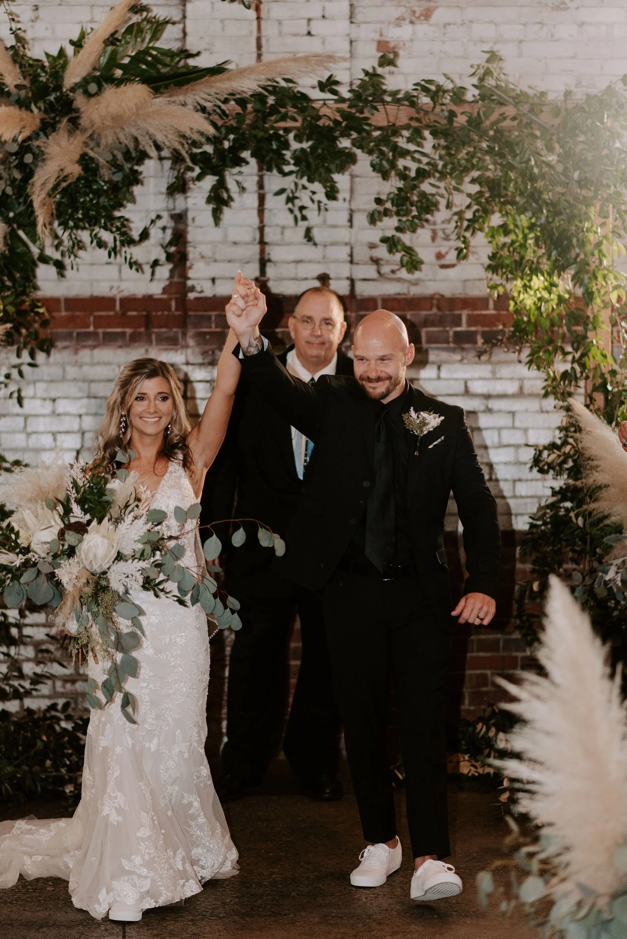 A newlywed couple celebrating their wedding with a dance or walk, holding hands up, surrounded by greenery and floral arrangements, with a brick wall background and a man officiant or witness smiling behind them.
